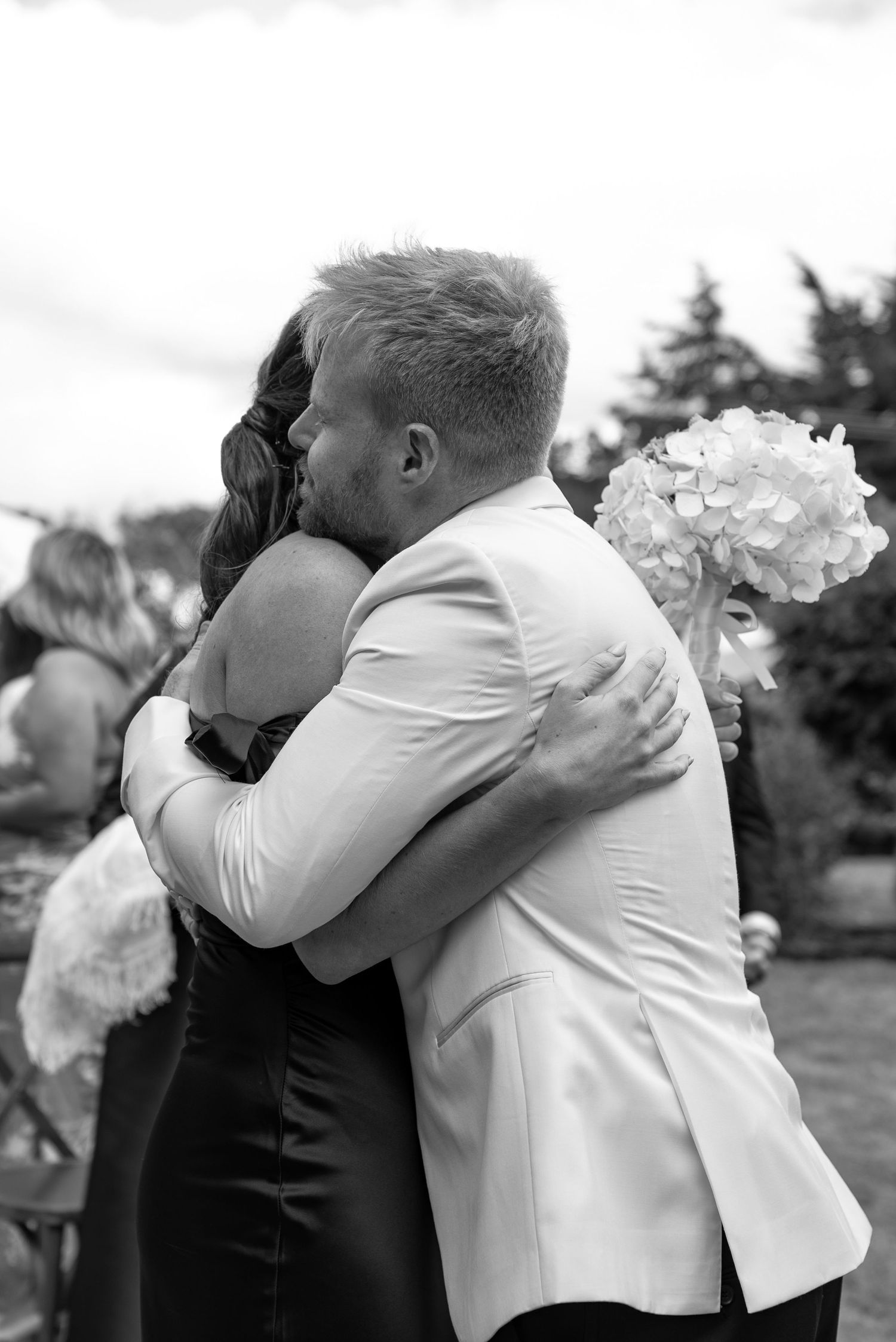 Black and white photo of a couple embracing at a wedding, groom in white jacket holding a bouquet of hydrangeas.