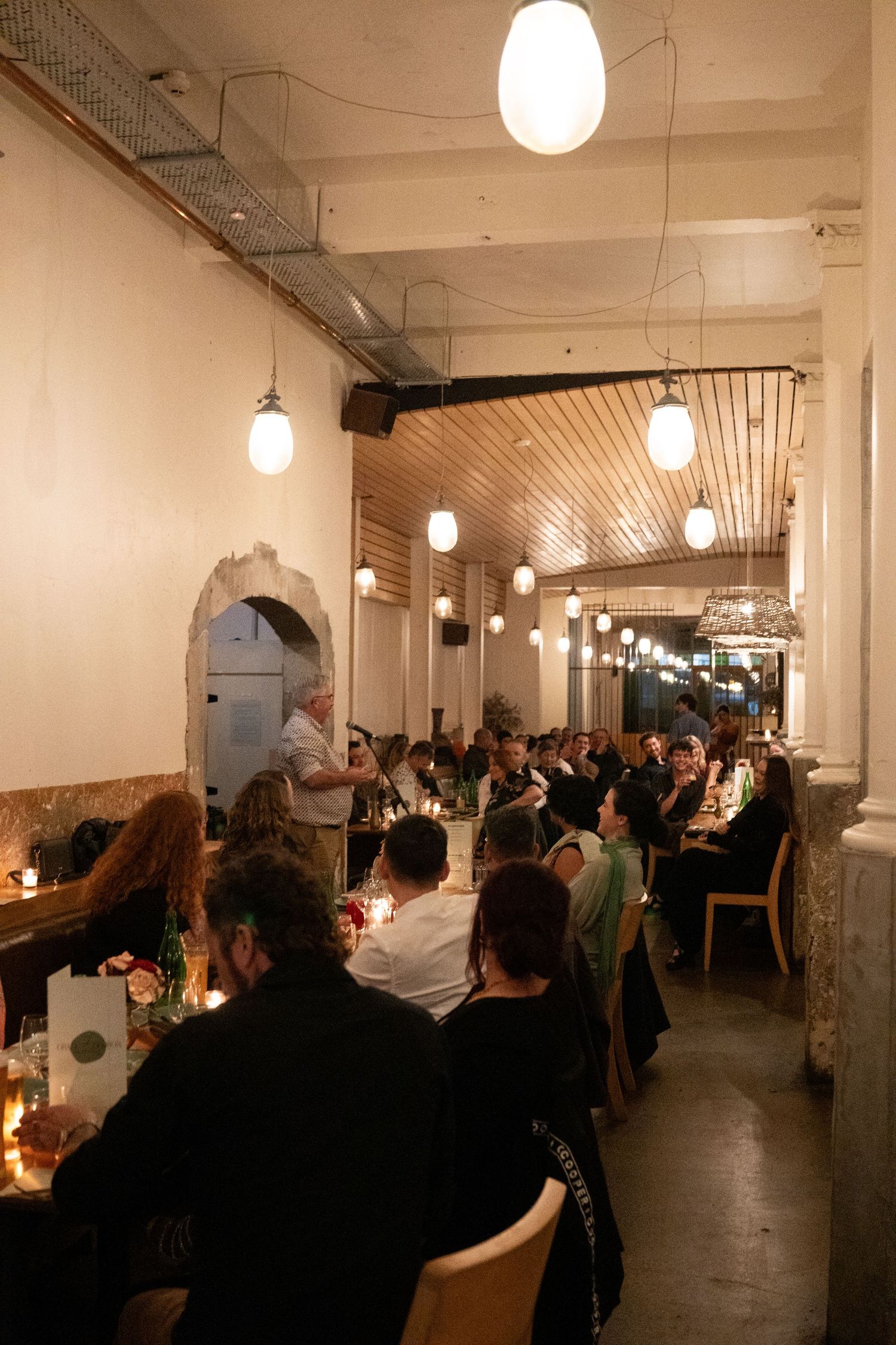 Guests dining at a long candlelit table in a warmly lit restaurant with pendant lights and wooden ceiling.