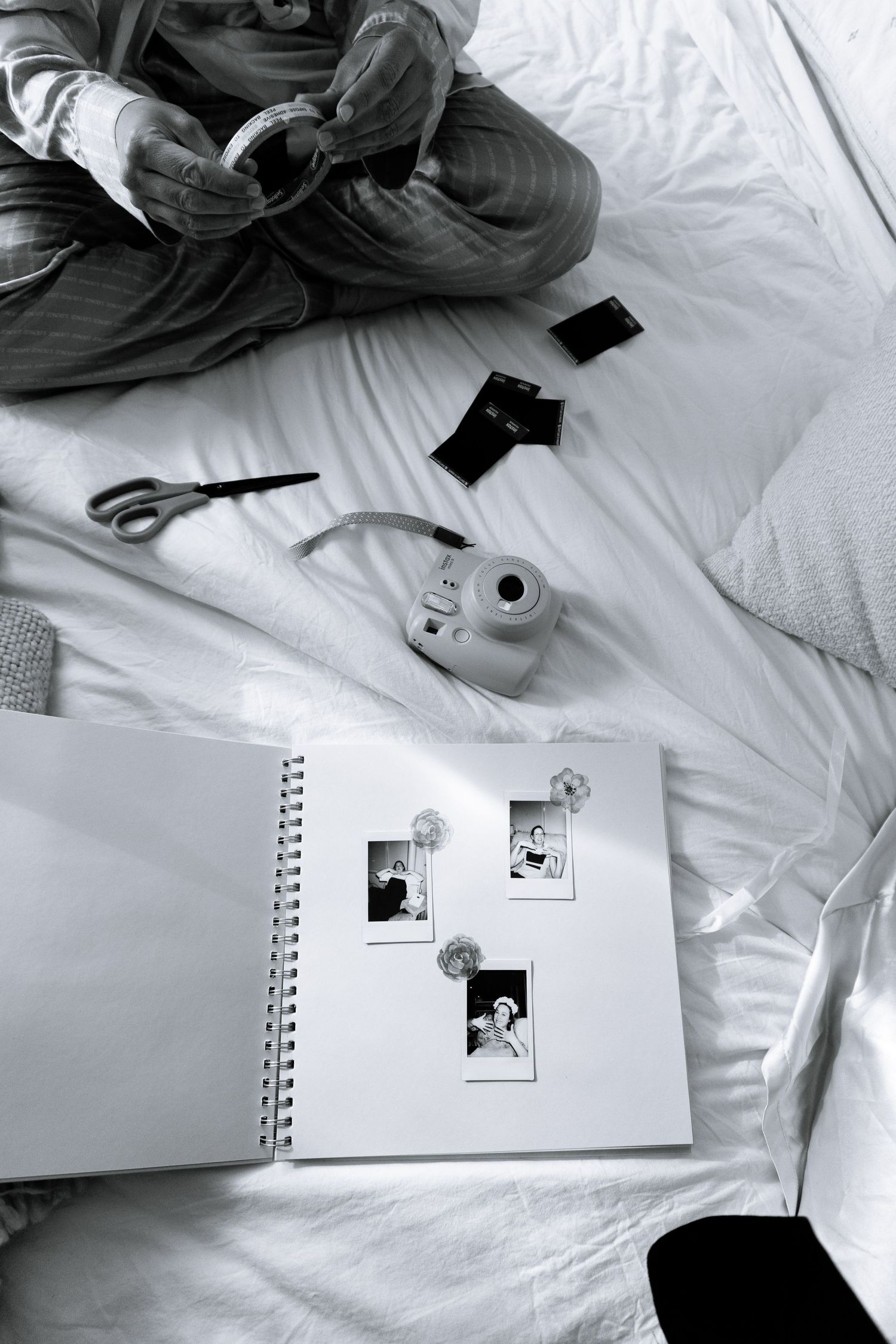 Black and white photo of a scrapbook with instant photos on a bed with a Polaroid camera, scissors, and film negatives.