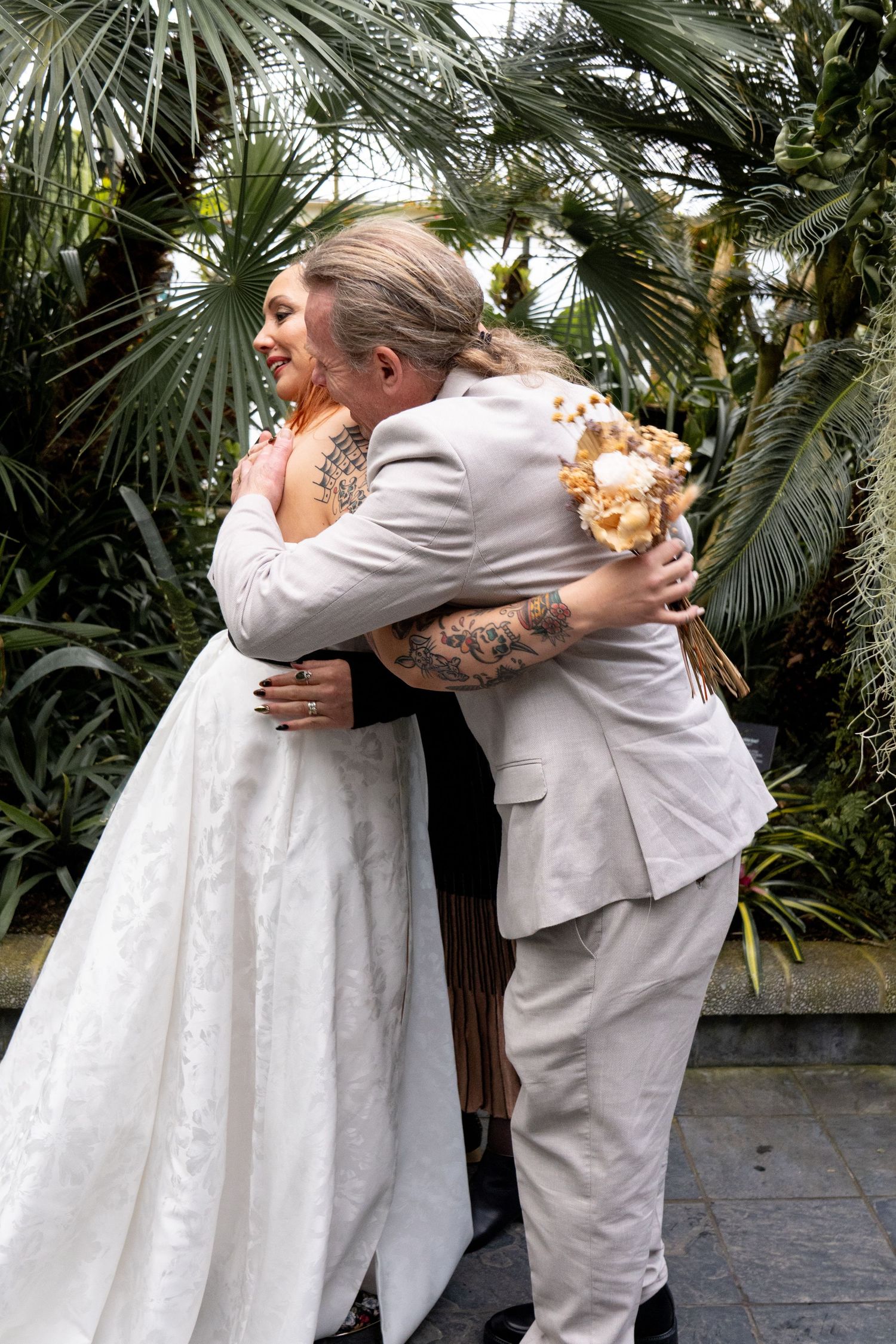 Bride and groom share a warm embrace outdoors surrounded by lush tropical greenery on their wedding day.