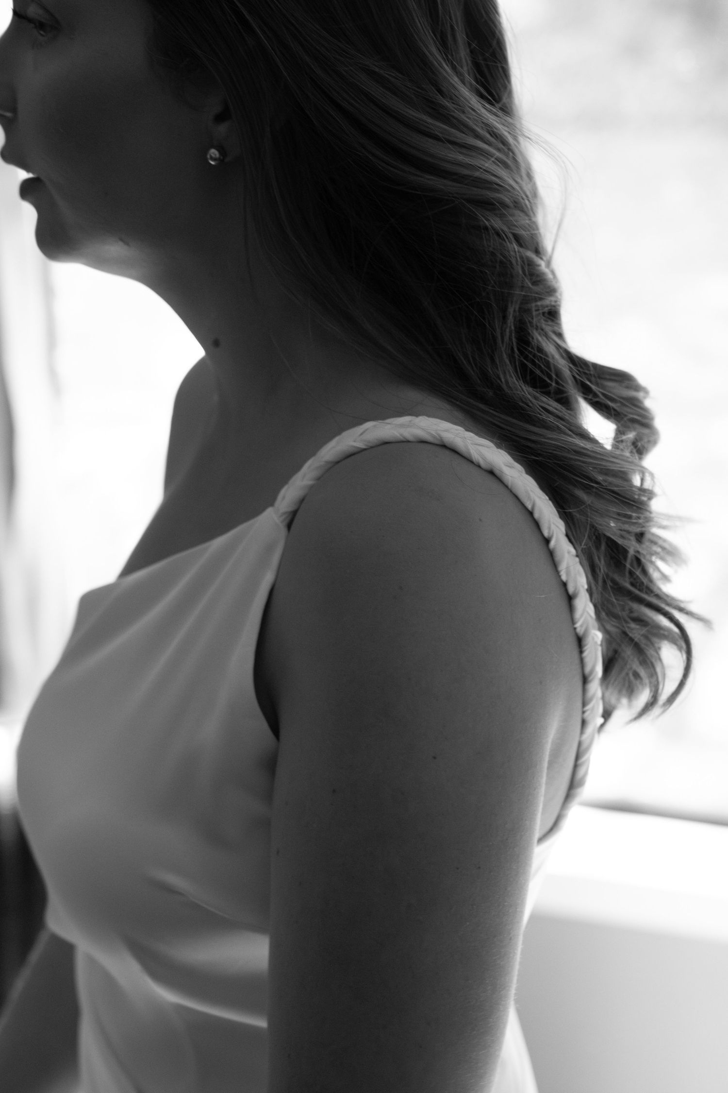 Black and white profile photo of a bride in a white dress with braided strap, long wavy hair, and pearl earring.