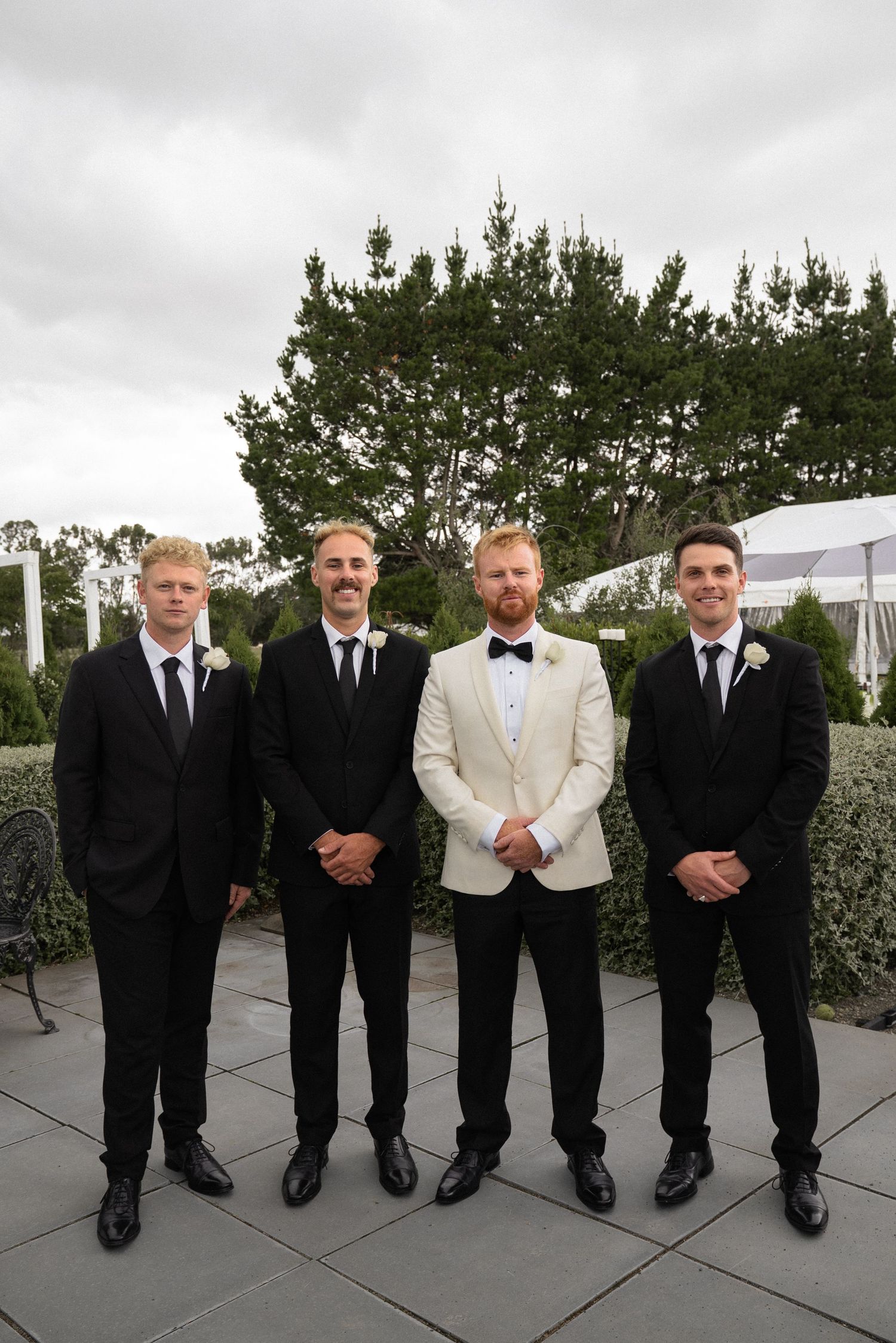 Groom in white tuxedo jacket stands with three groomsmen in black suits outdoors at a wedding venue.
