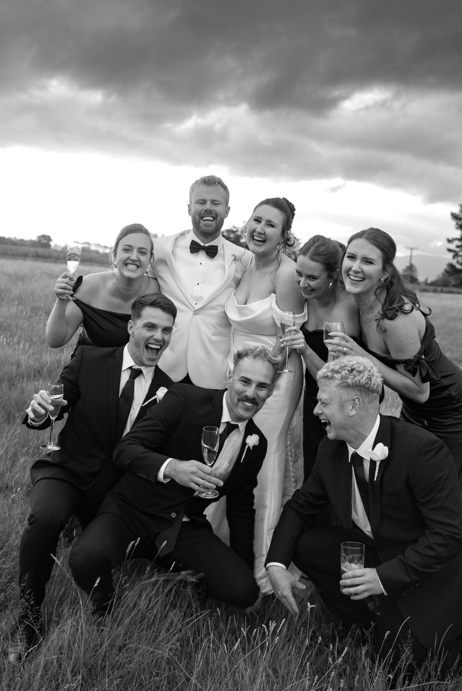 Black and white wedding party photo of bride, groom, and six friends laughing outdoors with champagne glasses.
