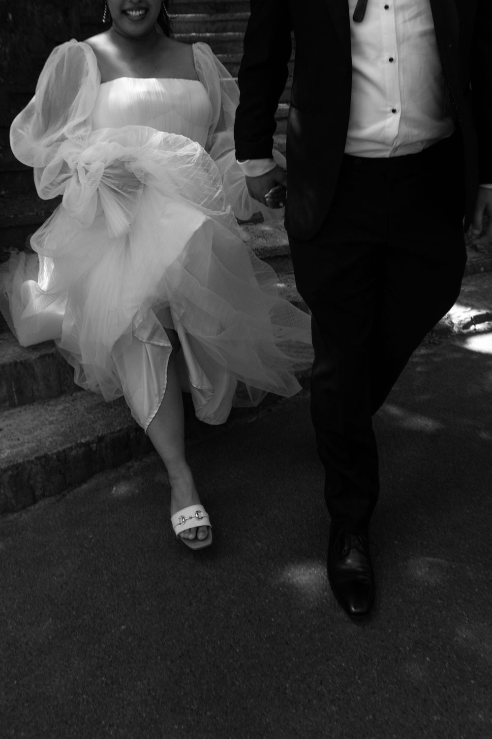 Black and white photo of bride in tulle gown and groom in tuxedo walking down steps together.