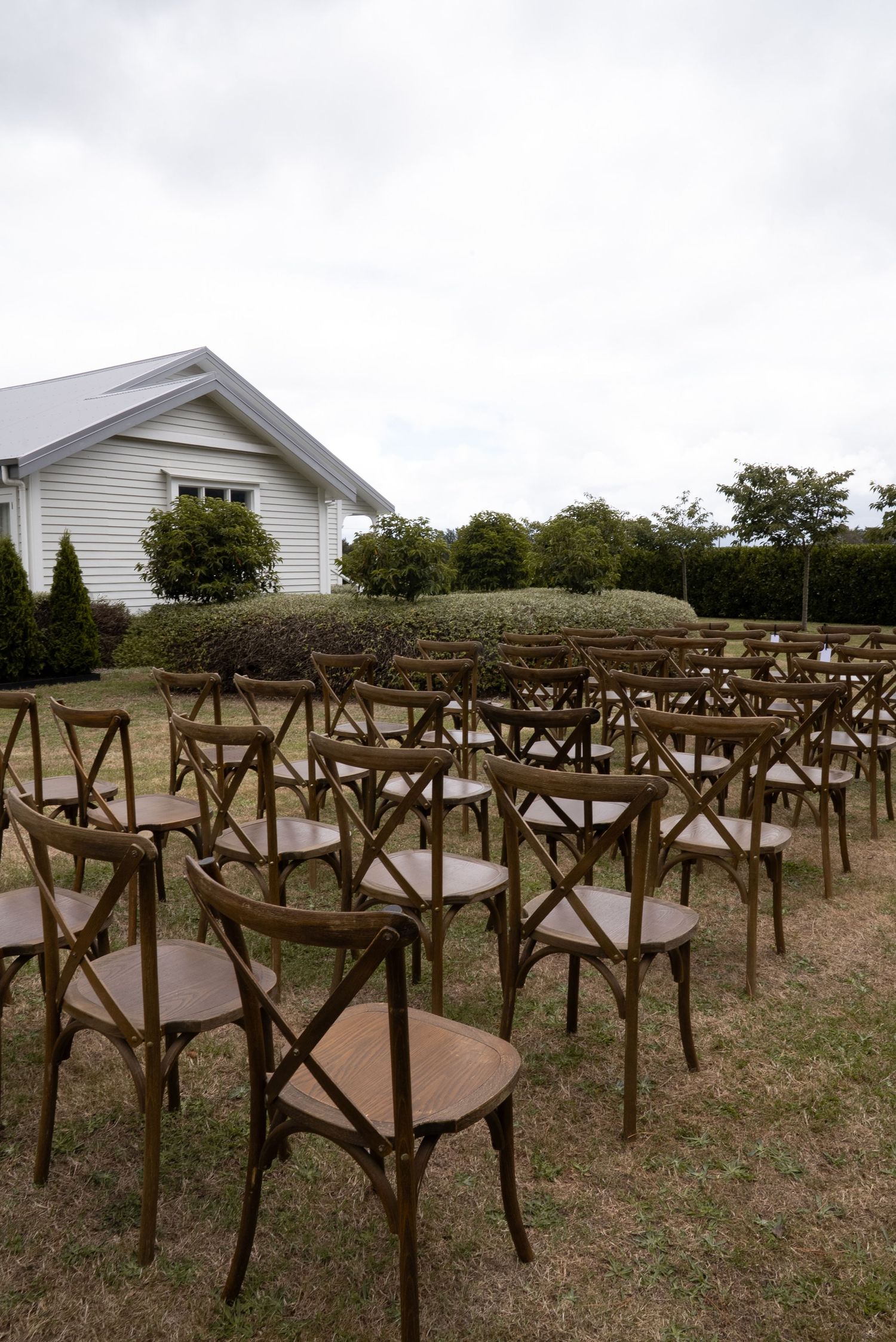 Rows of rustic wooden cross-back chairs arranged outdoors on grass for a garden wedding ceremony near a white building.