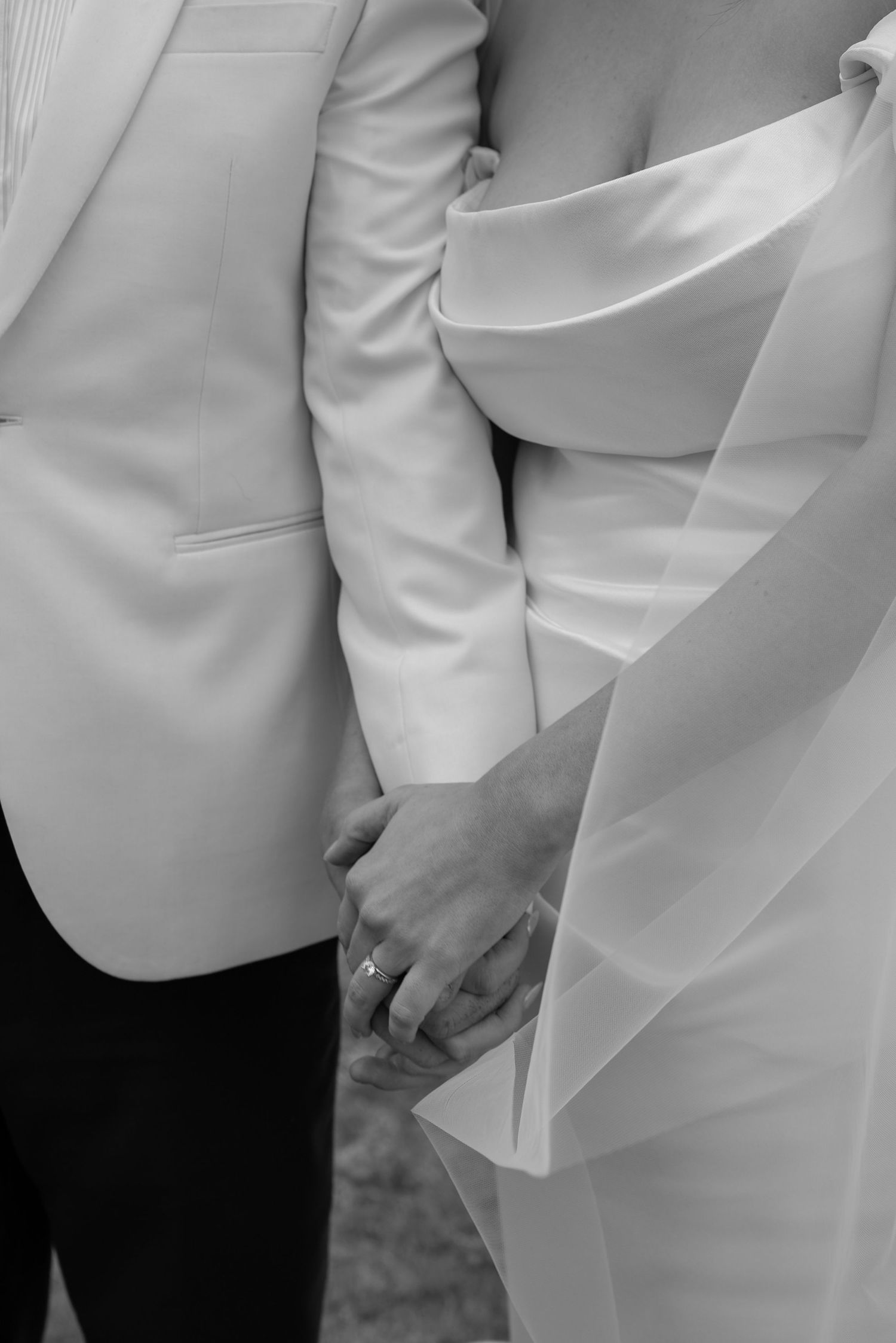 Black and white photo of a bride and groom holding hands, showcasing wedding rings and elegant white attire.