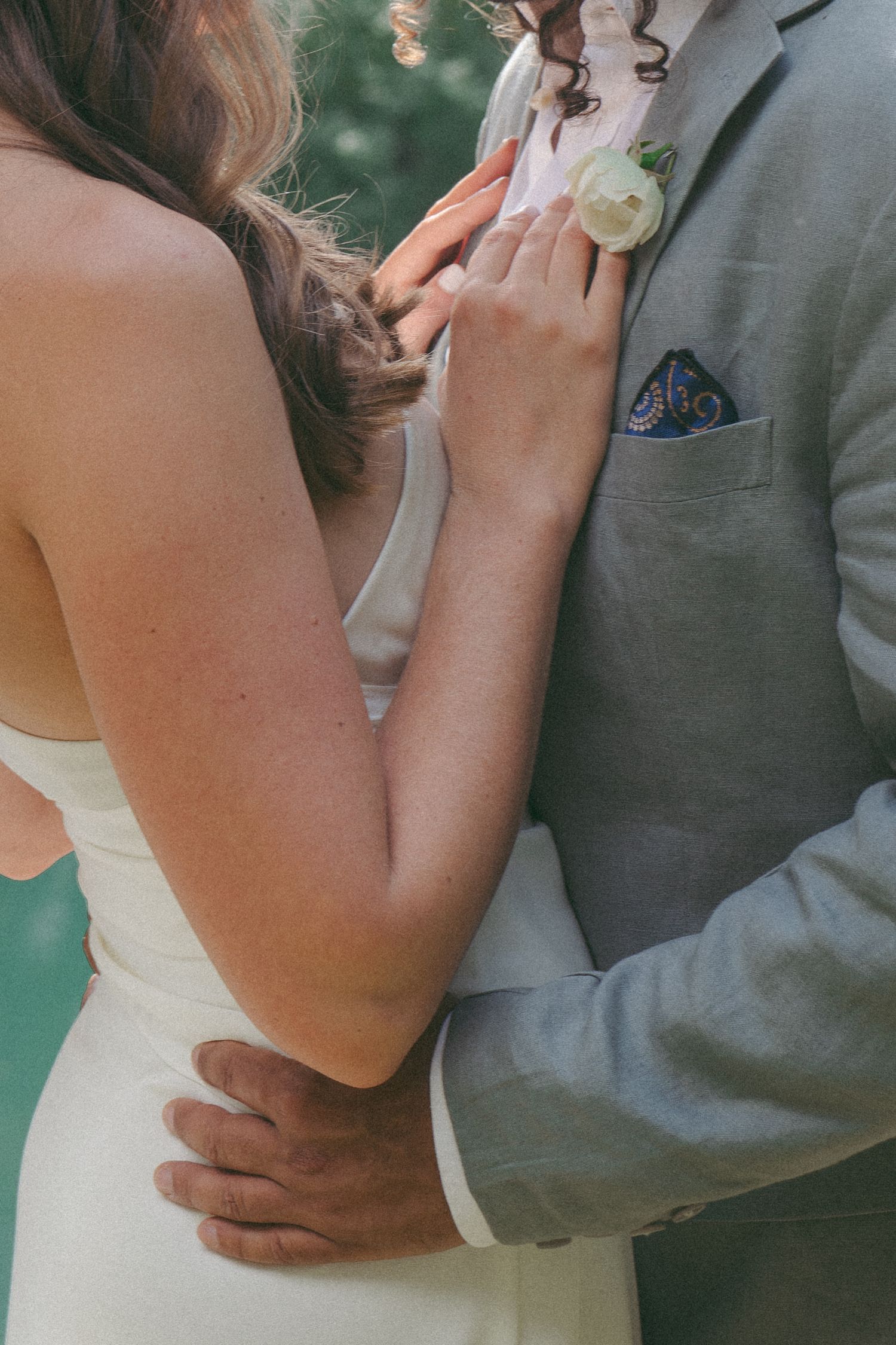 A bride in white dress and groom in gray suit with boutonniere embrace closely at a wedding ceremony.