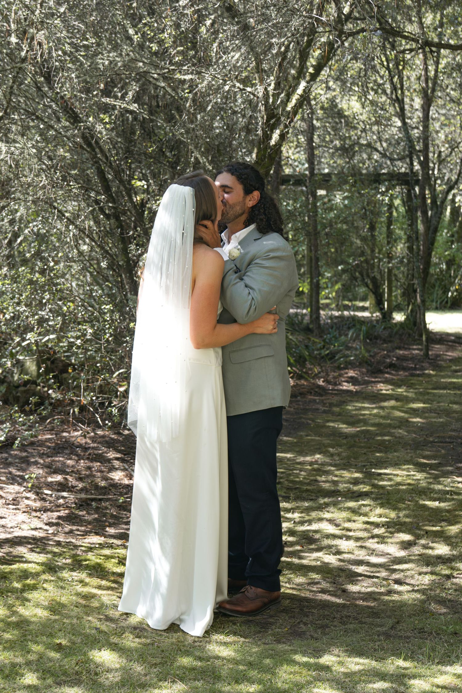 Bride and groom share first kiss outdoors in wooded garden setting, bride in white gown and veil, groom in gray suit.