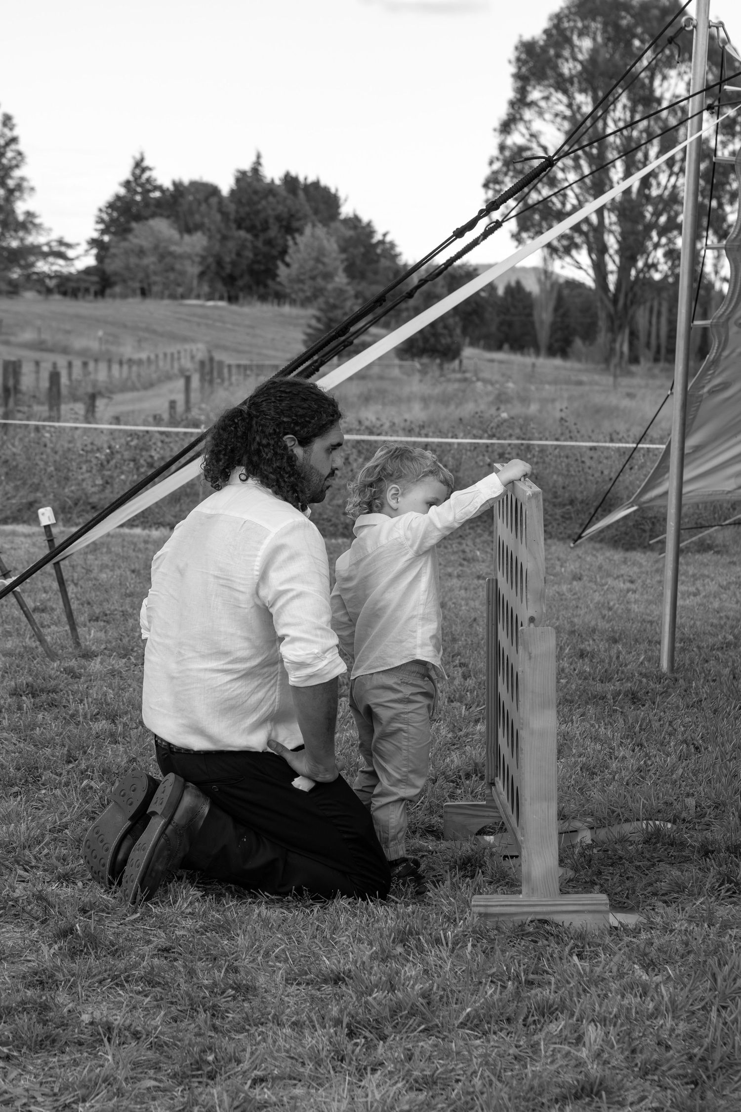 Black and white photo of a parent kneeling behind a toddler exploring a wooden structure in a rural field.