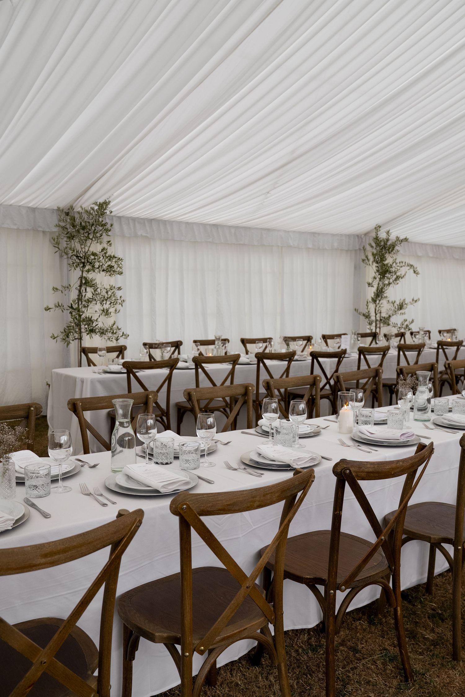 Elegant marquee wedding reception with rustic cross-back chairs, white linen tables, glassware, and greenery decor.