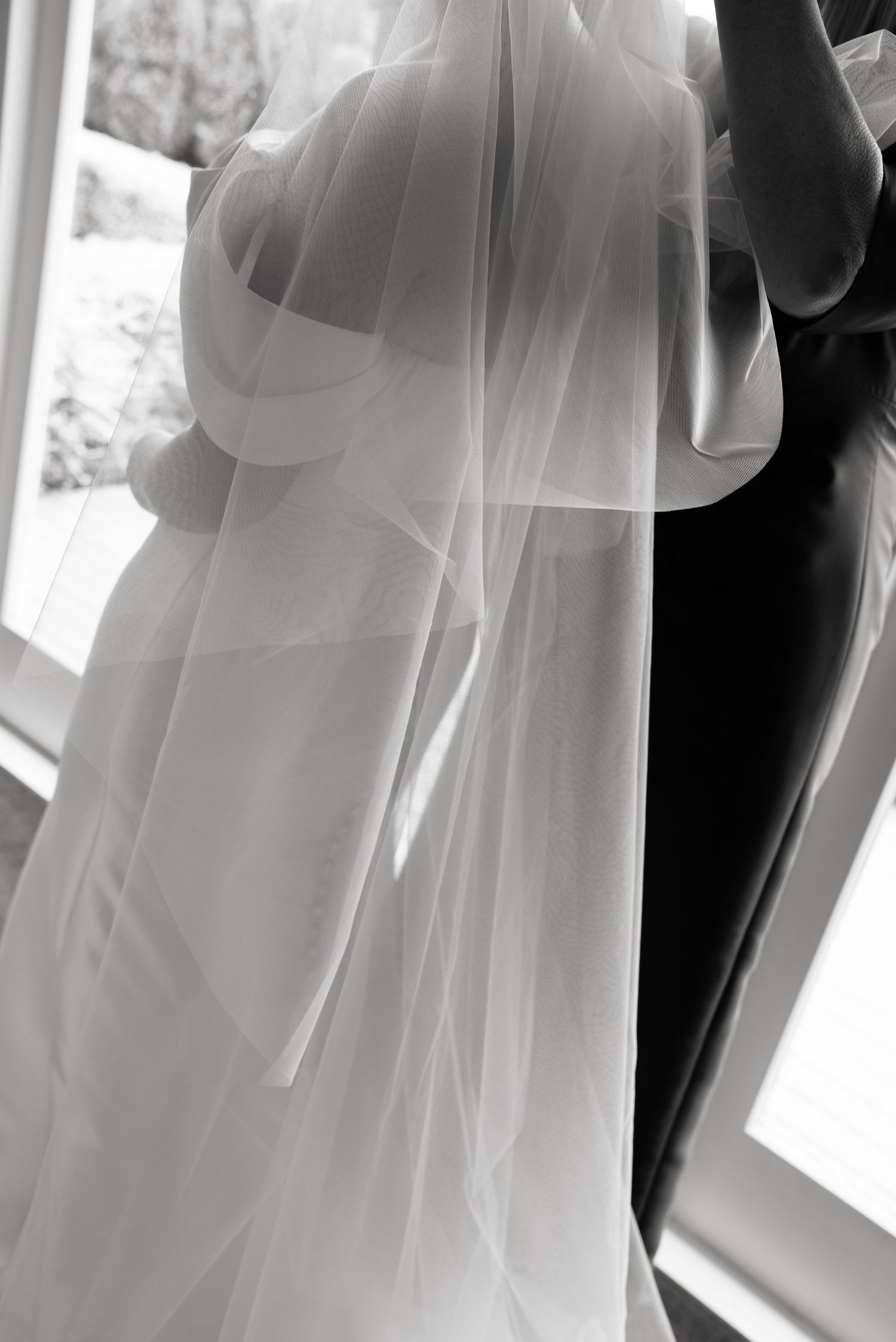Black and white photo of a bride in a flowing wedding dress and veil being helped by an assistant near a window.