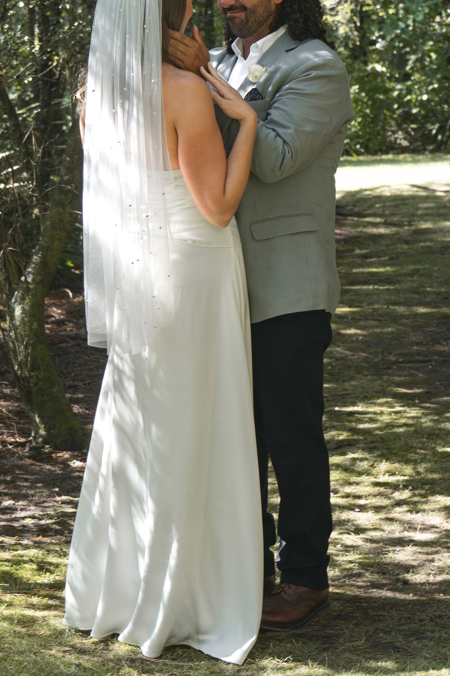 Bride in white gown and veil embraces groom in gray suit outdoors in a sunlit forest setting.