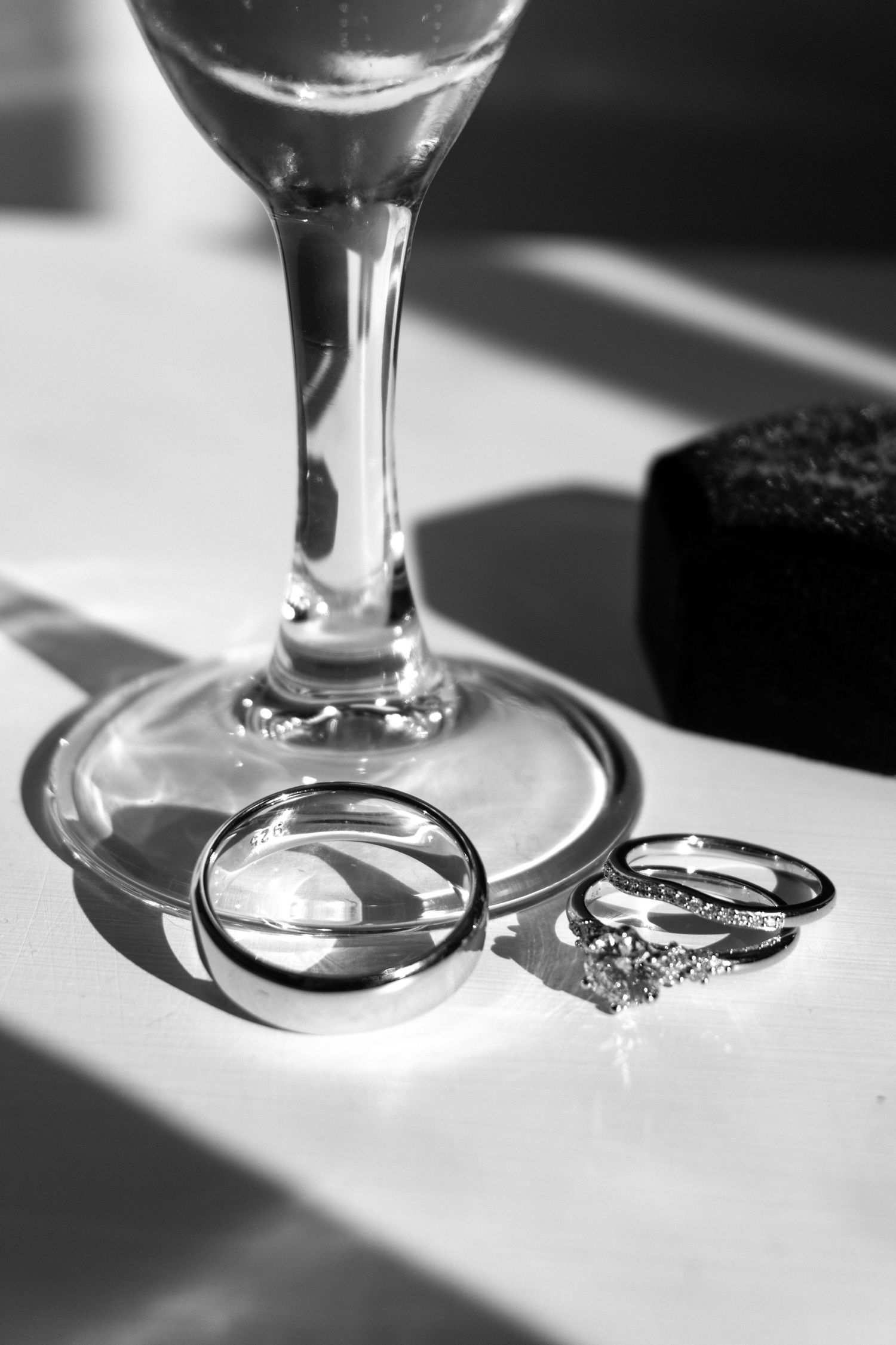 Black and white photo of wedding rings and engagement ring beside a champagne glass on a white surface.