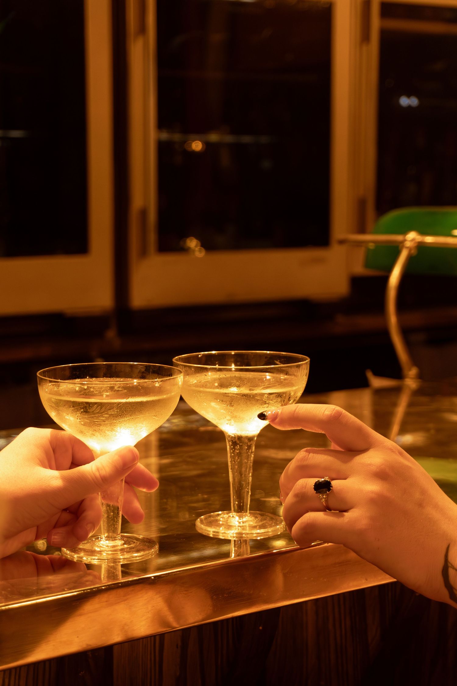 Two hands clinking coupe champagne glasses on a golden bar top in a warmly lit elegant setting.