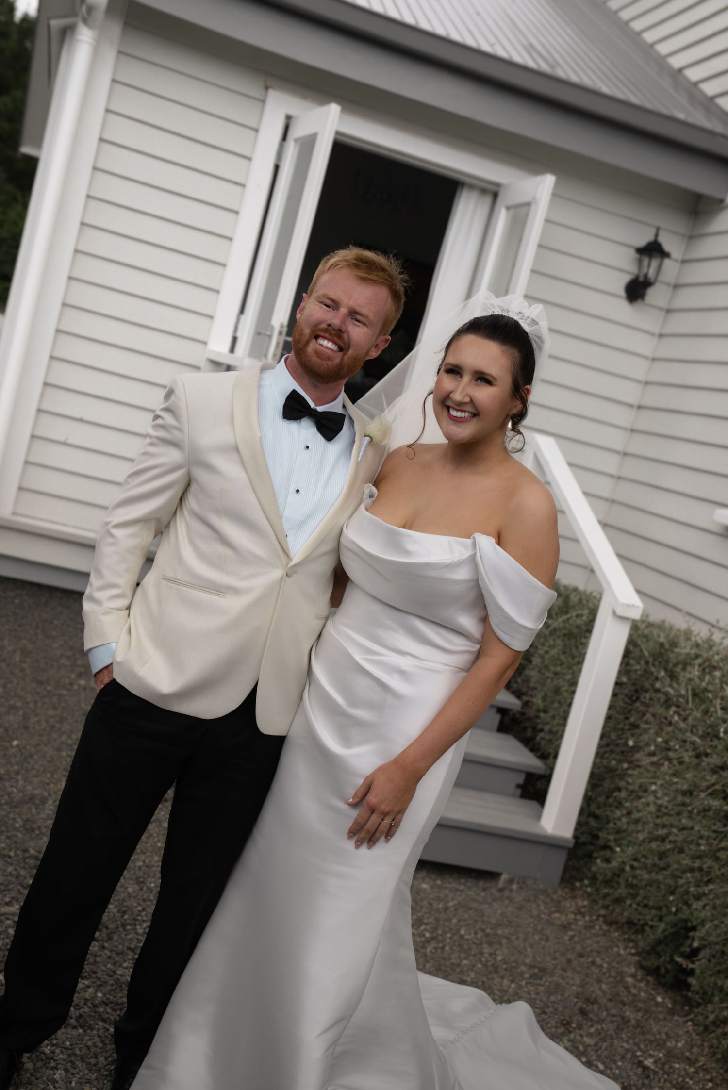 A smiling bride and groom pose in wedding attire outside a white chapel, she in an off-shoulder gown, he in a cream tuxedo.