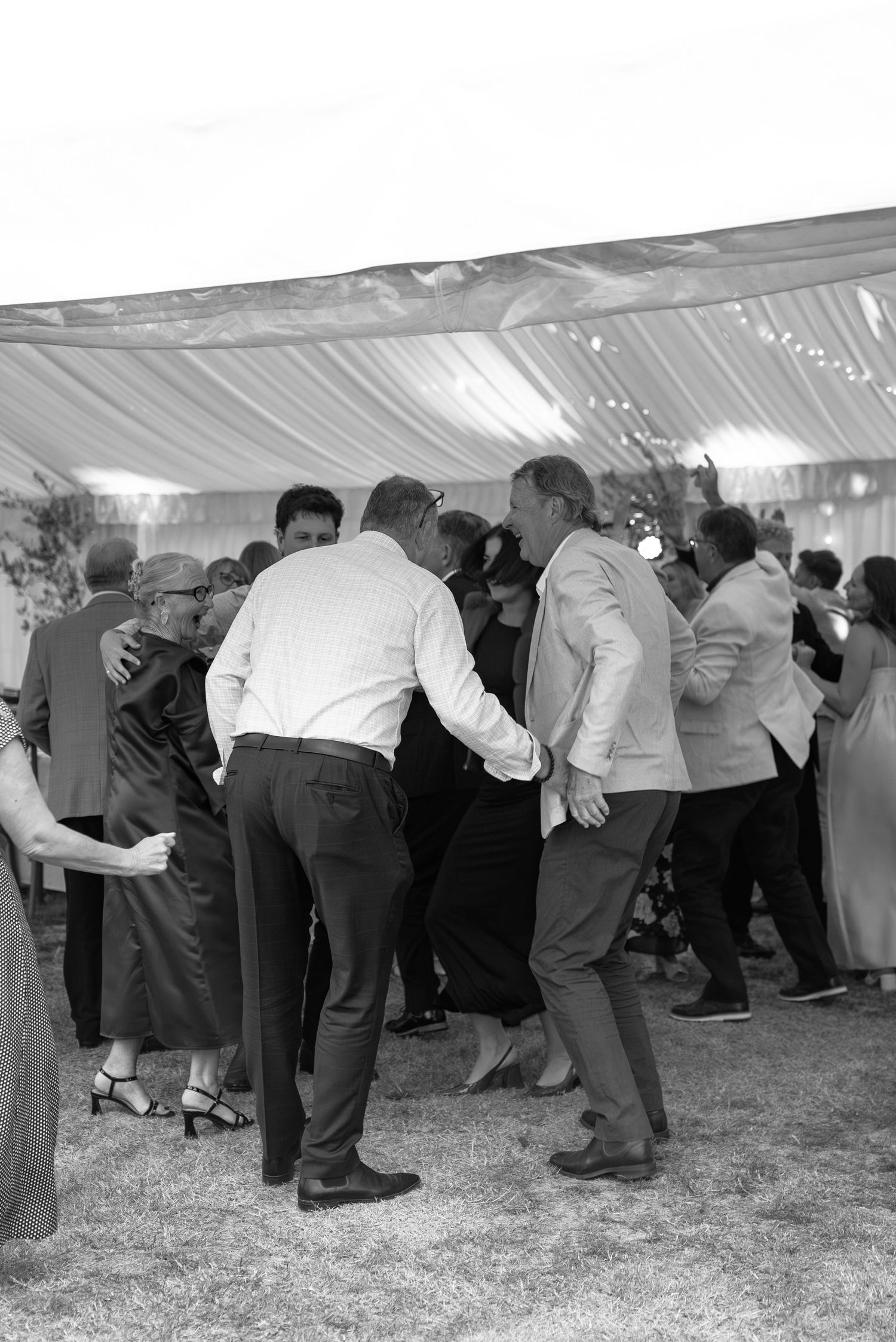 Black and white photo of elegantly dressed guests dancing together under a draped tent at a wedding reception.