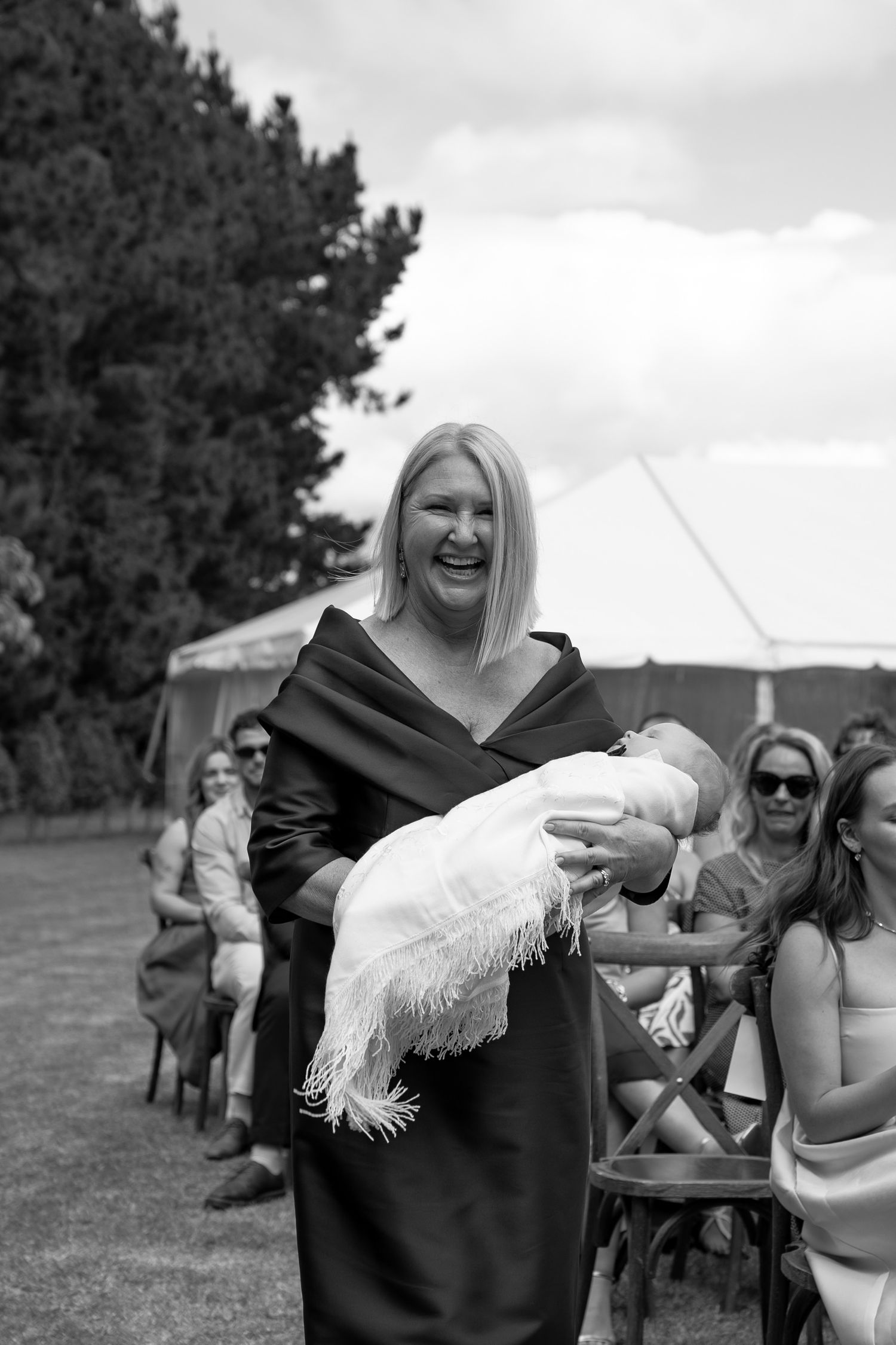Woman in black dress joyfully carries a baby in white christening gown outdoors at a garden ceremony.