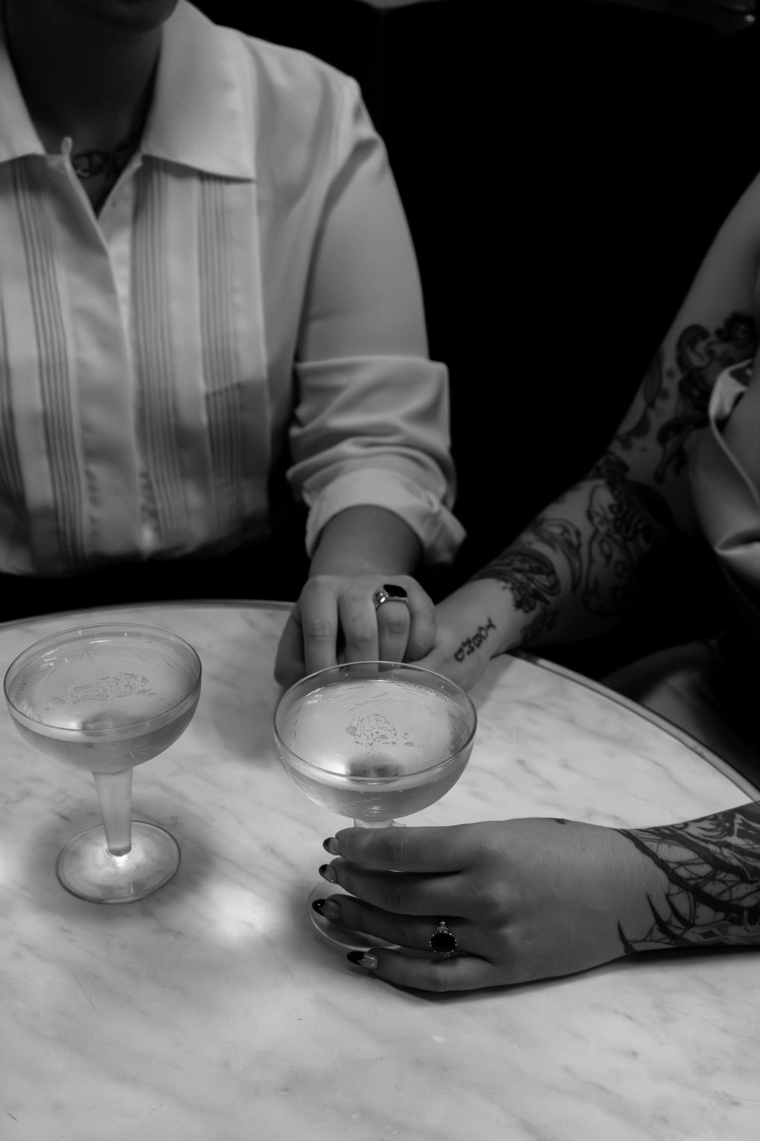 Two people sit at a marble table holding frothy cocktails, one with tattooed hands and dark nails, in black and white.