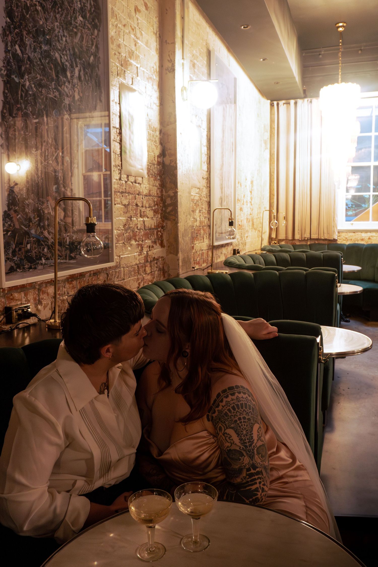 A bride and groom share a romantic kiss in an elegant restaurant booth, with cocktail glasses on the table.