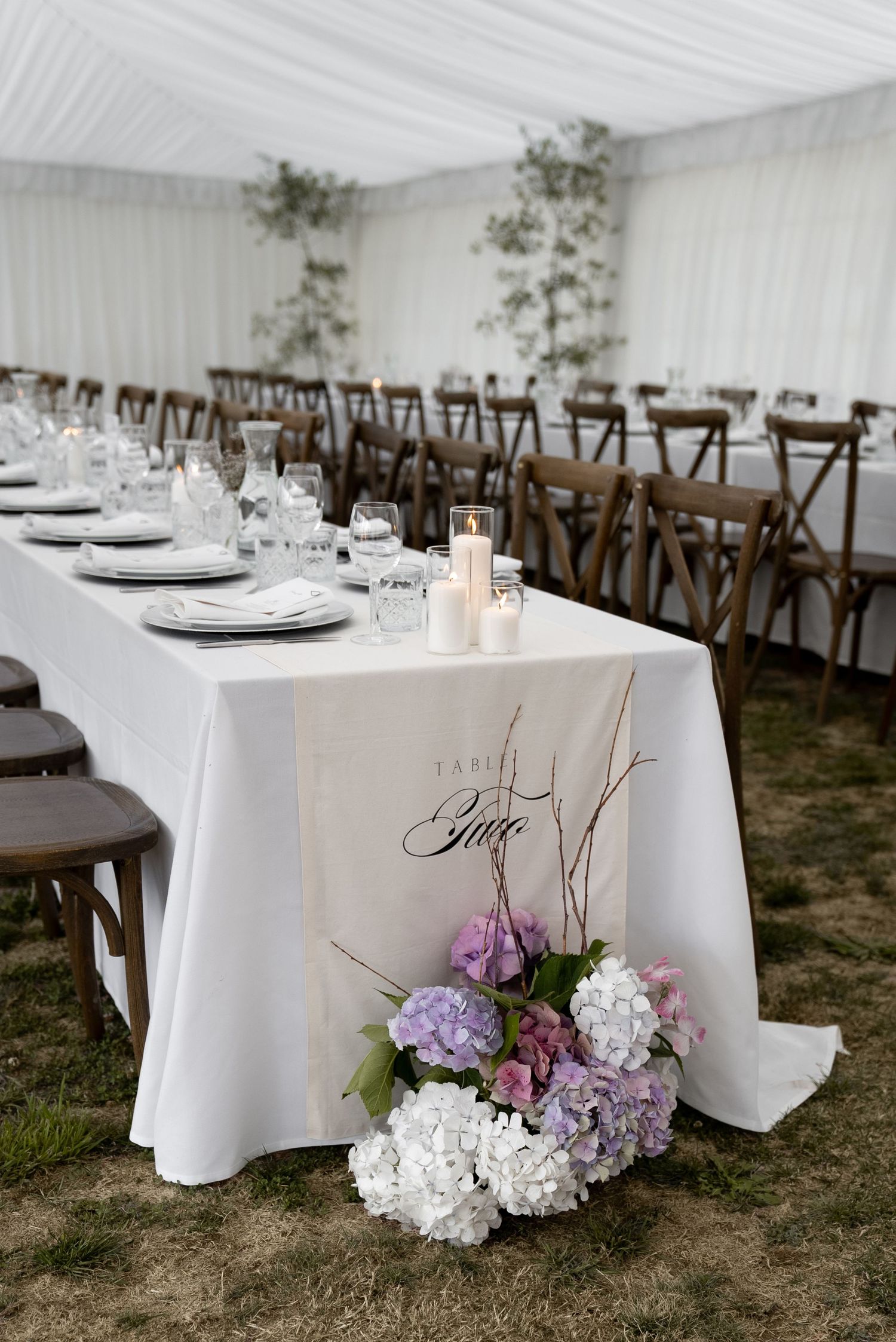 Elegant wedding reception tent with long white-draped tables, hydrangea floral arrangements, candles, and rustic cross-back chairs.
