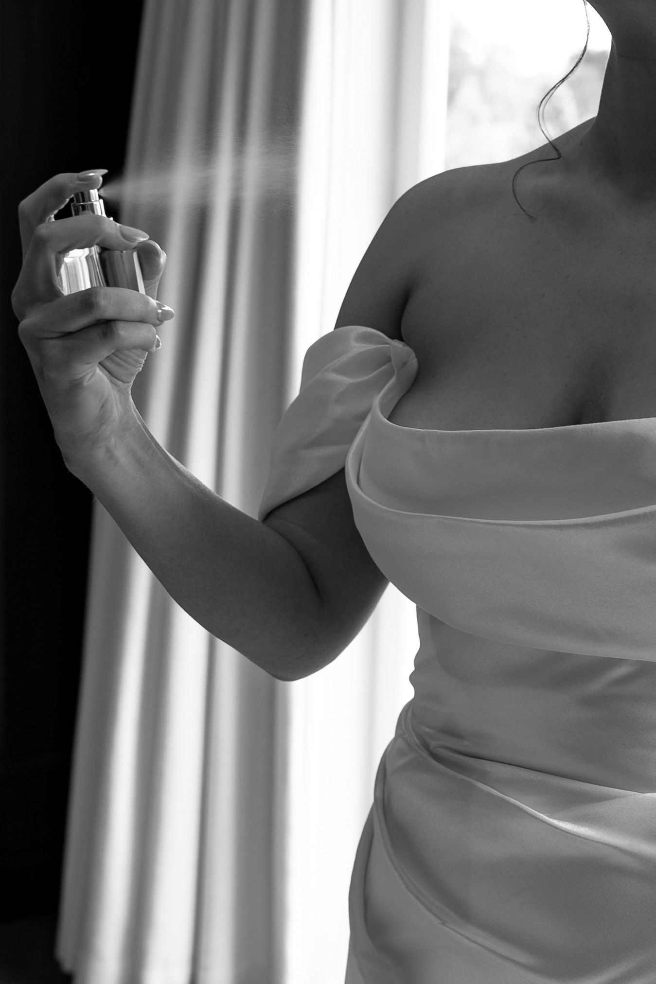 Black and white photo of a woman in an off-shoulder dress spraying perfume, elegant bridal getting ready moment.