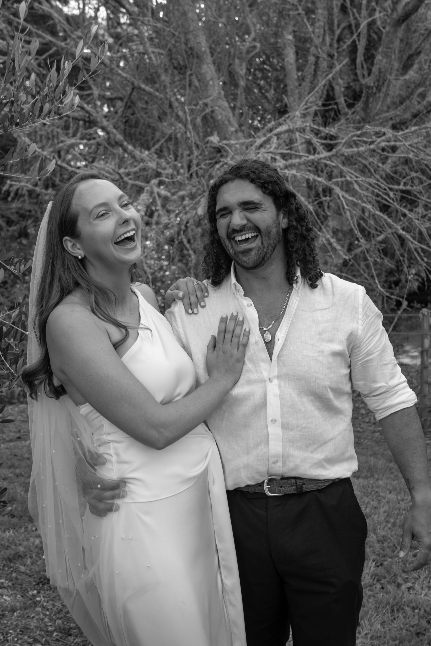 Black and white photo of a laughing bride in white dress and groom in white shirt sharing a joyful moment outdoors.