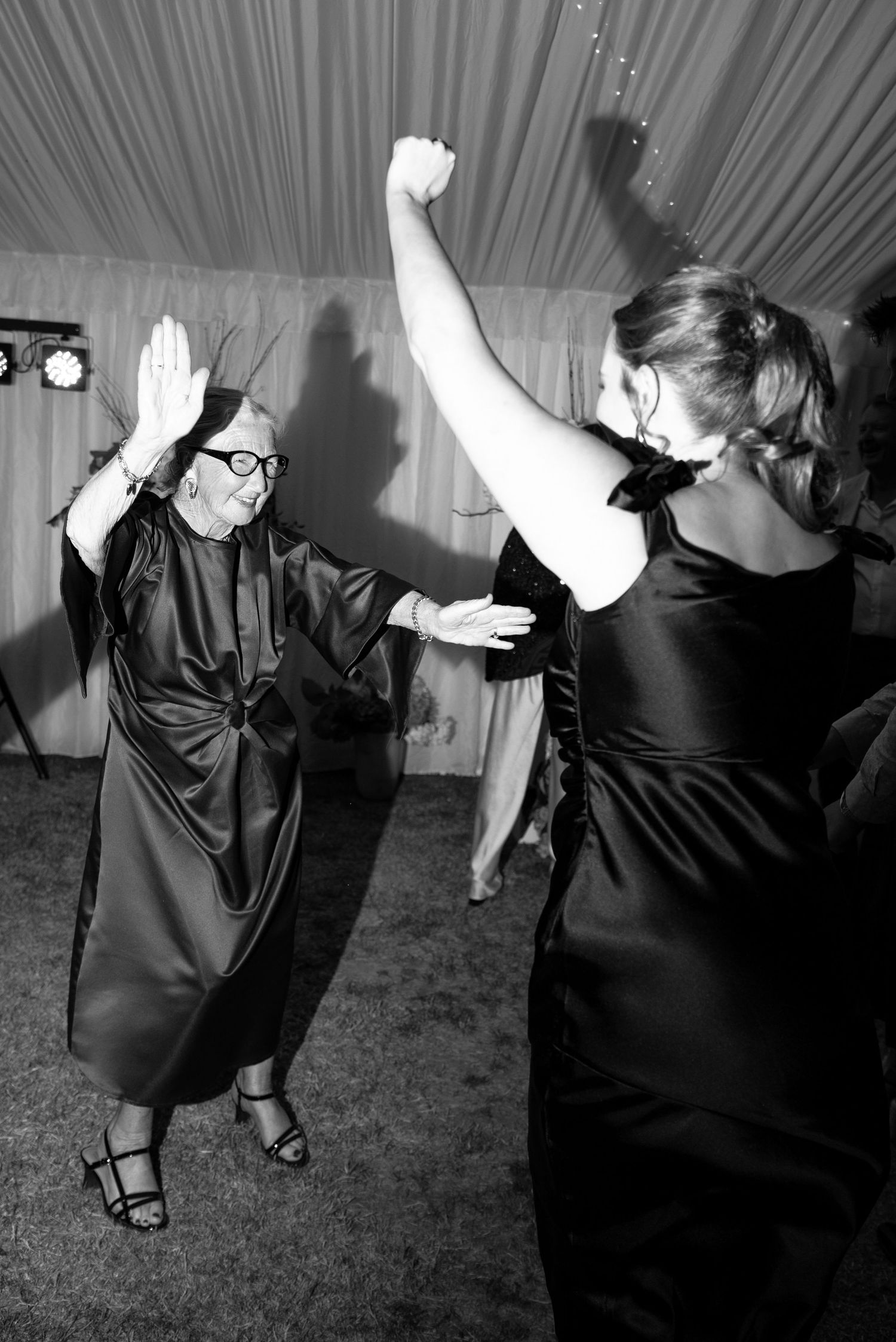 Two women dancing joyfully at a formal event, raising their arms in a black and white photo taken inside a tent.