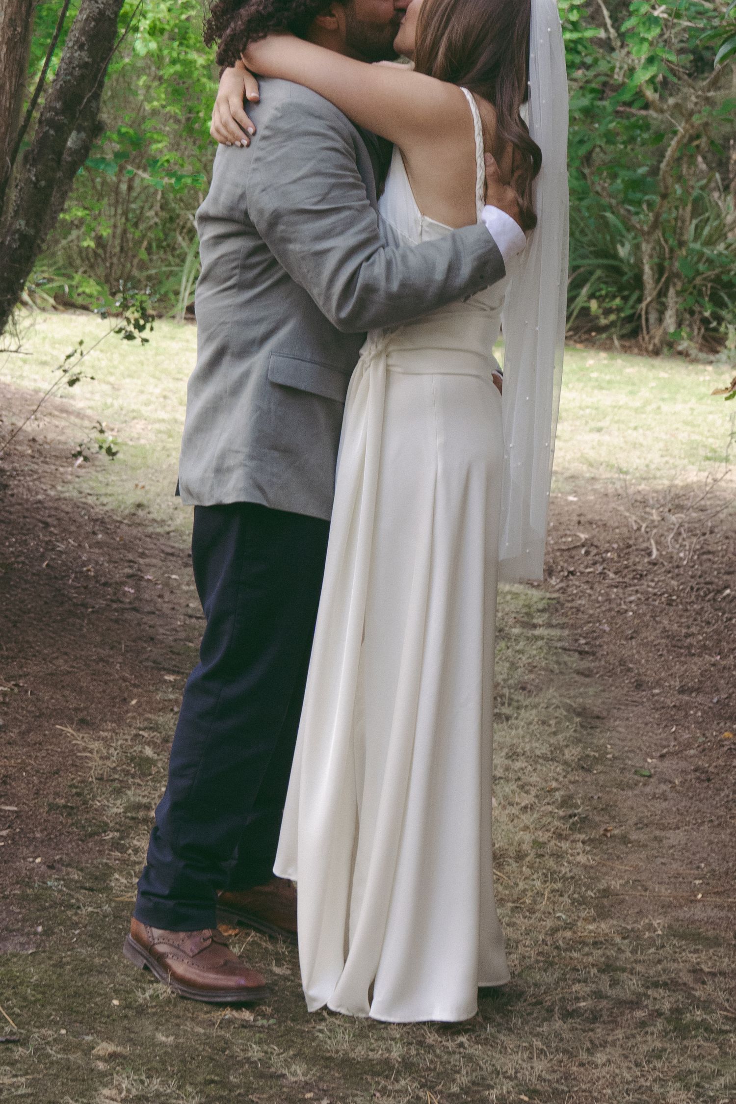 A couple shares a romantic kiss outdoors, bride in white gown with veil, groom in gray blazer and navy pants.