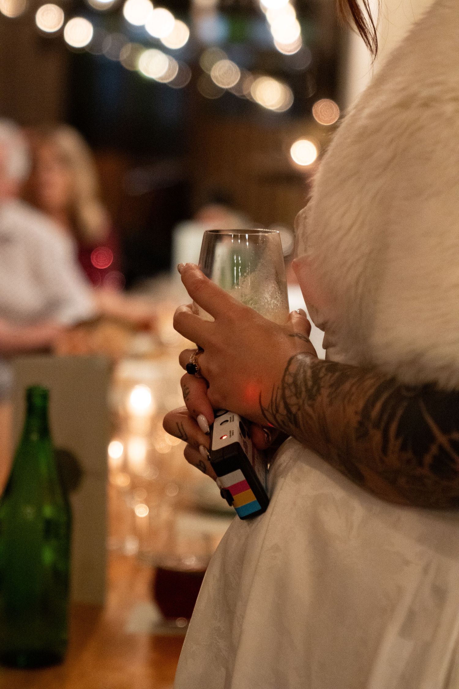 Tattooed hands holding a champagne glass at a candlelit dinner party with bokeh string lights.