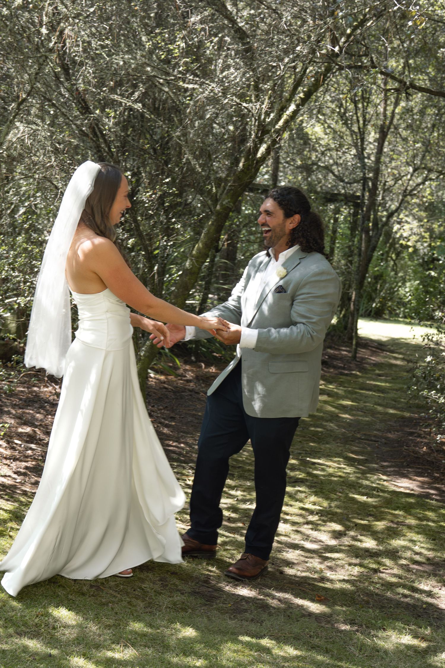 Bride and groom holding hands and laughing together on a sunlit forest path, bride in white gown with veil.