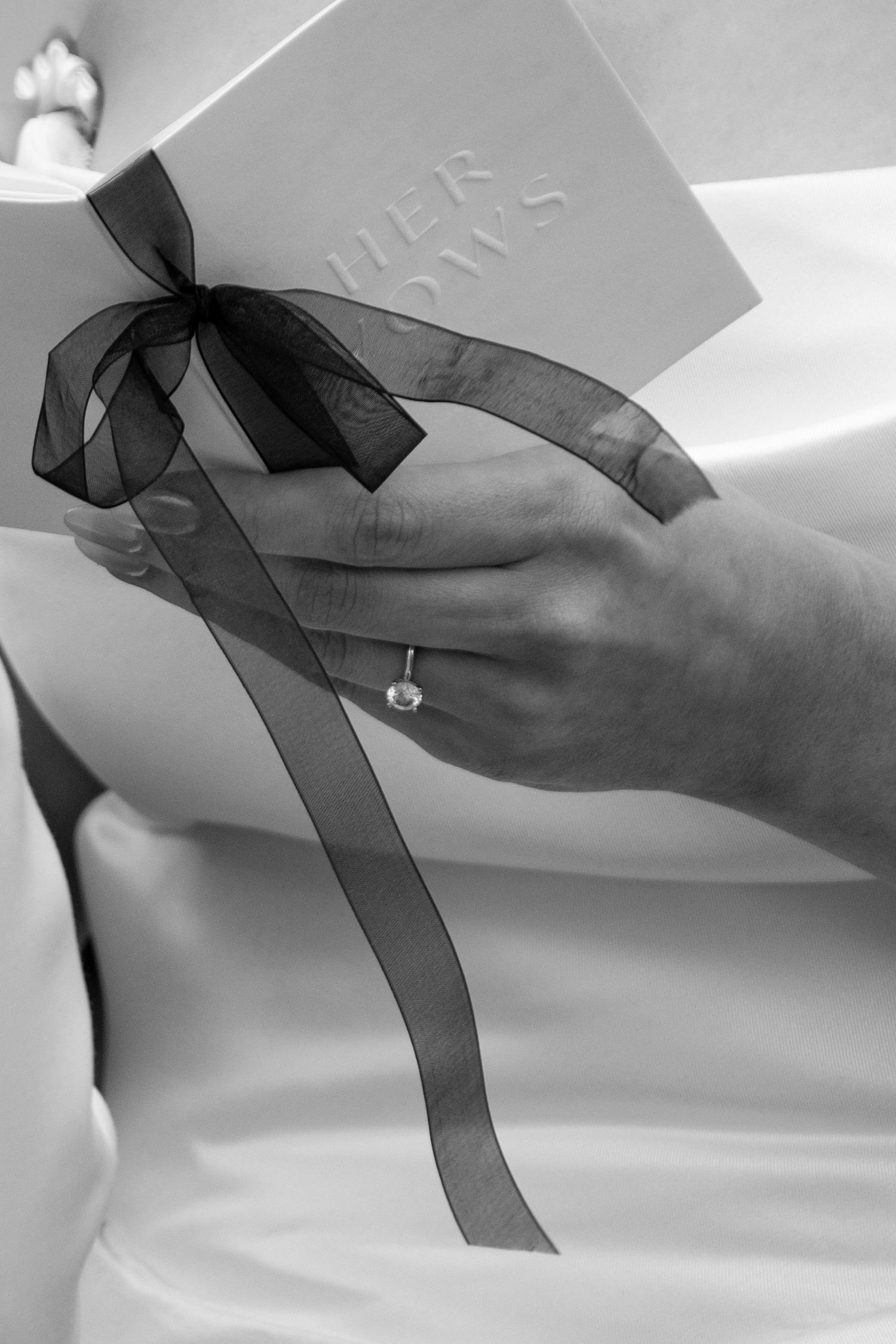 Bride holding her vows booklet with black ribbon bow, wearing engagement ring, black and white wedding photo.