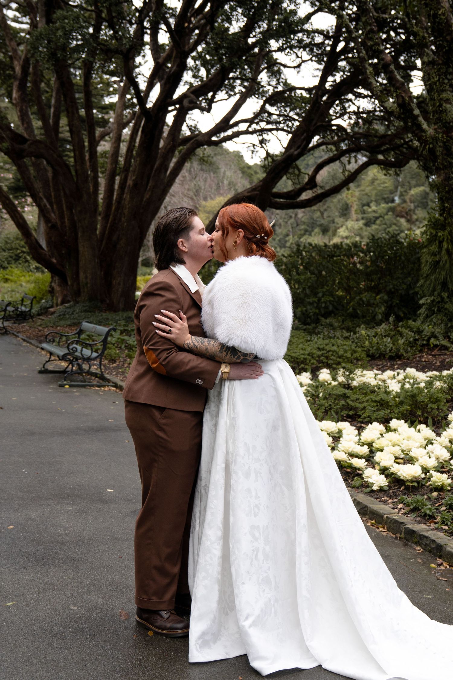 Couple sharing a romantic moment in a garden, one in a brown suit and one in a white bridal gown with fur wrap.