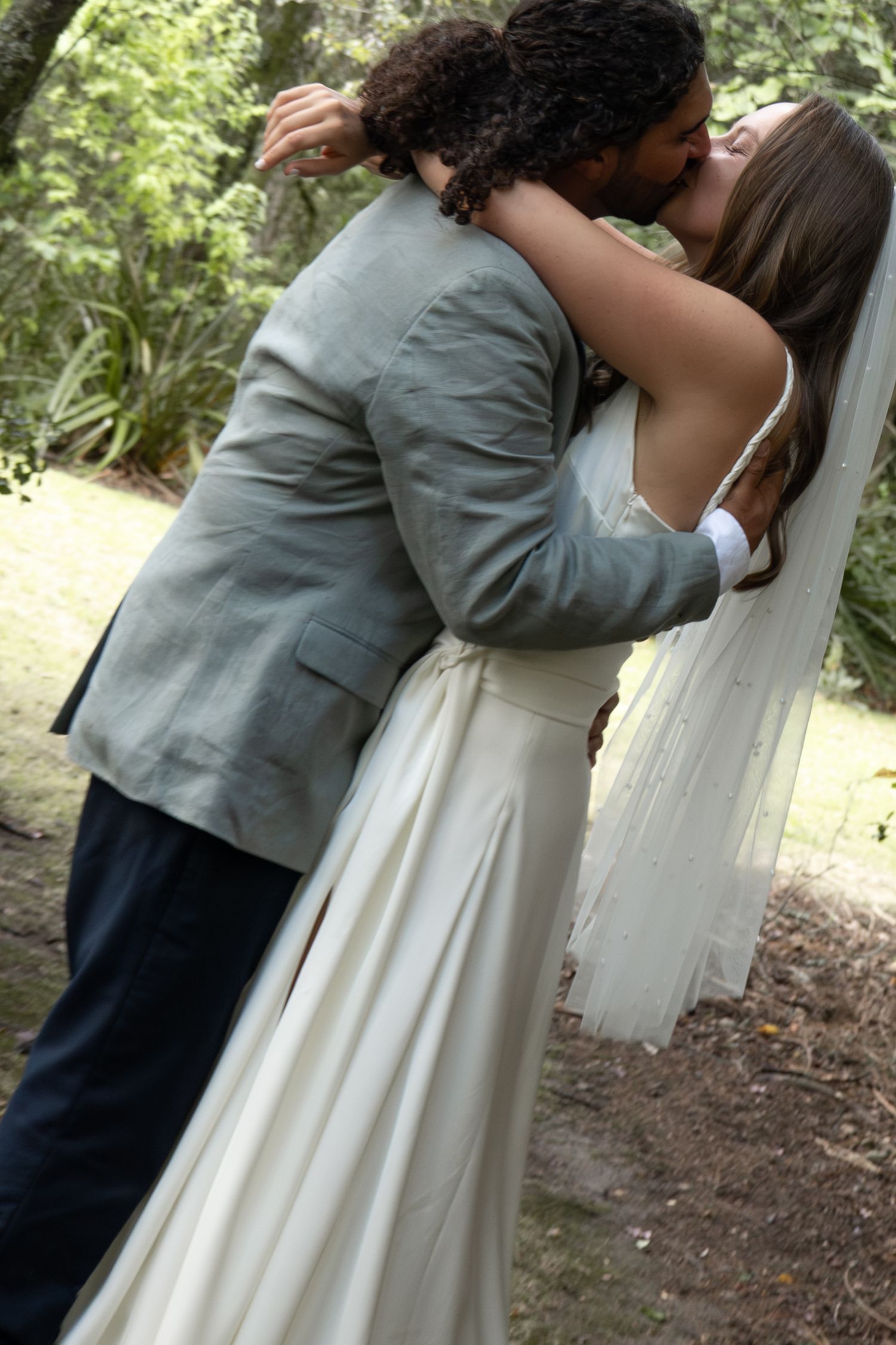 Bride and groom sharing a romantic kiss outdoors in a garden, wearing wedding dress with veil and gray suit.