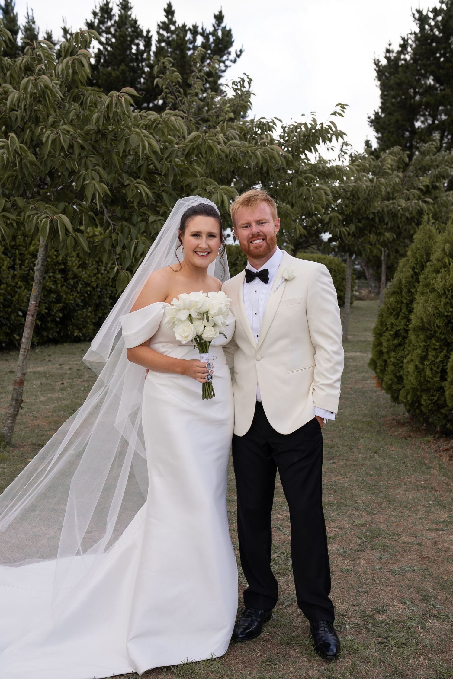 Bride in white gown with veil and groom in ivory tuxedo jacket pose together outdoors among green trees.