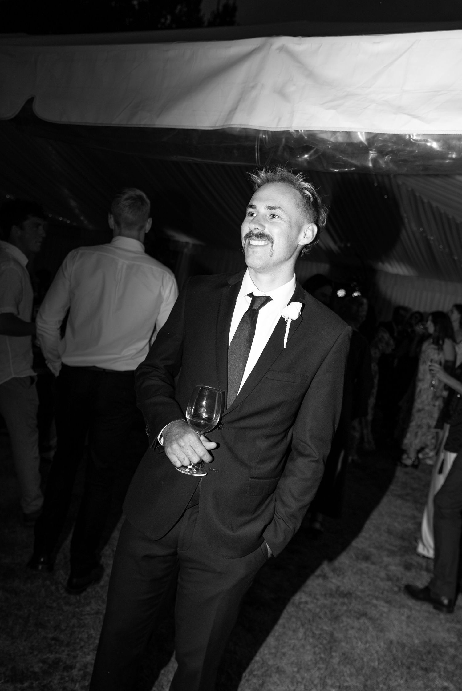 Man in suit holding wine glass smiling at outdoor evening wedding reception, black and white photo.