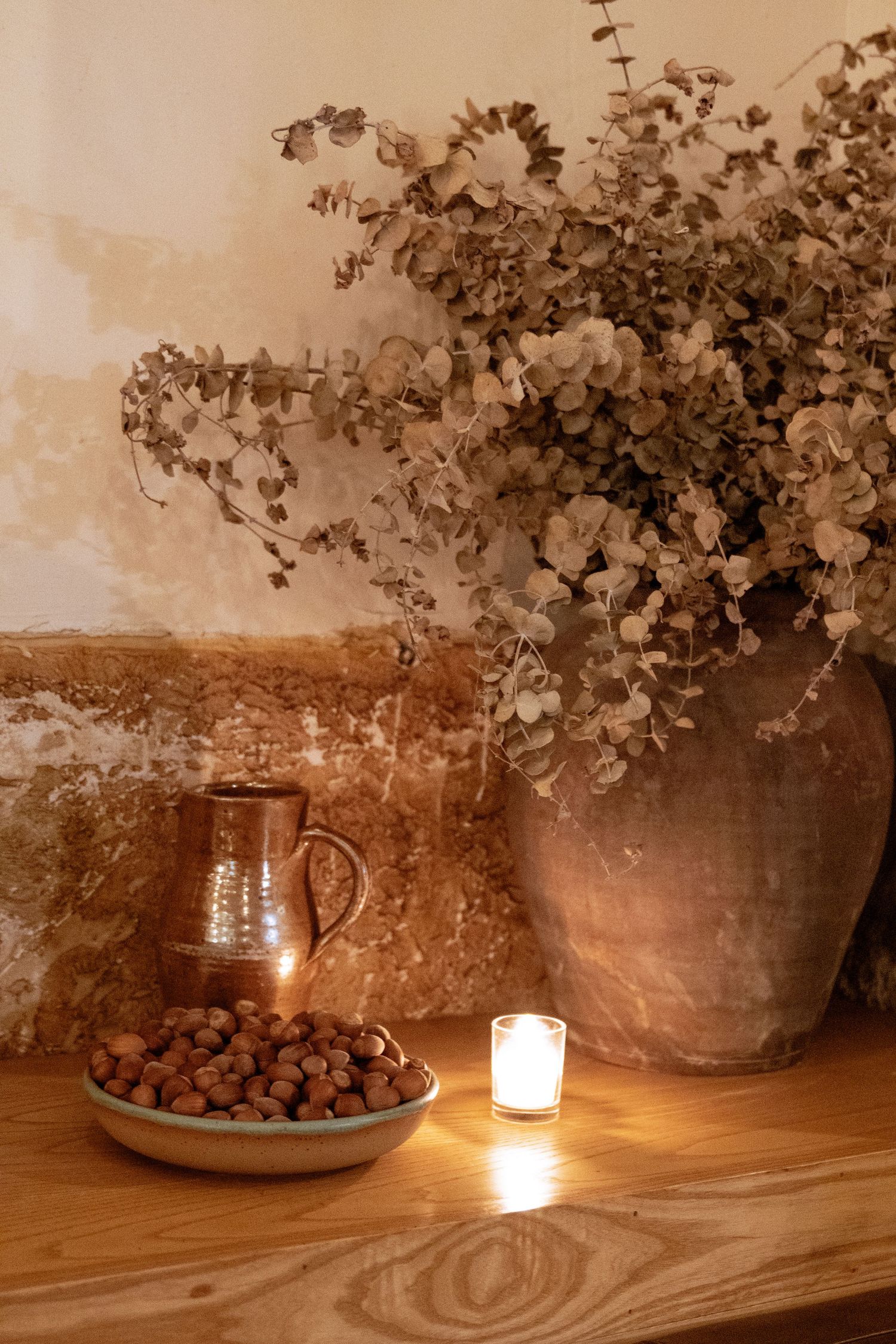 Rustic still life with dried eucalyptus in clay vase, hazelnuts in bowl, copper jug, and lit candle on wooden shelf.