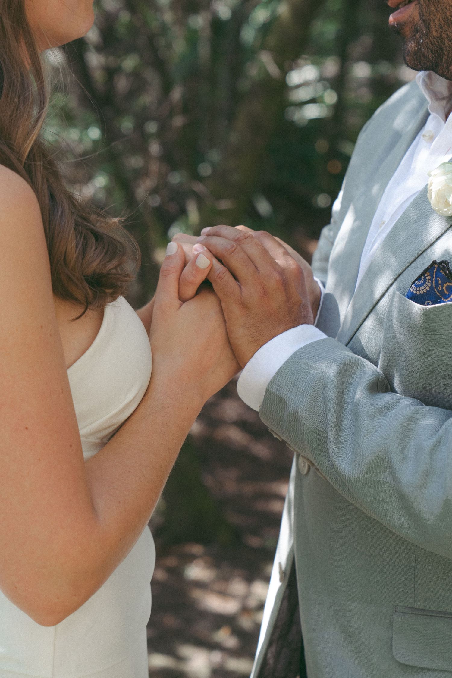 Bride and groom holding hands during outdoor wedding ceremony, groom wearing light gray suit with blue paisley pocket square.