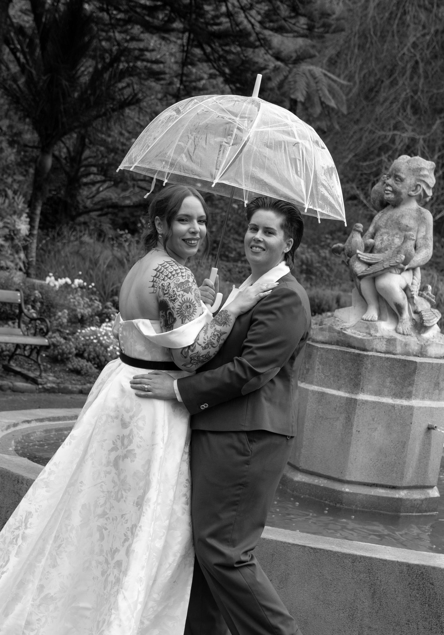 Black and white photo of two brides sharing an umbrella in the rain near a cherub fountain on their wedding day.