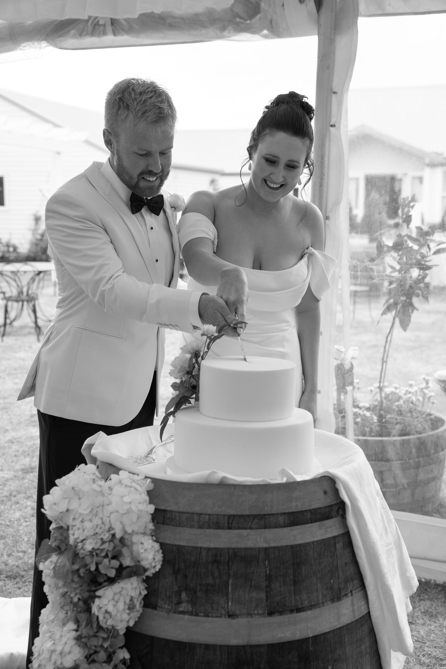 Bride and groom cutting their white tiered wedding cake on a barrel outdoors in black and white photo.