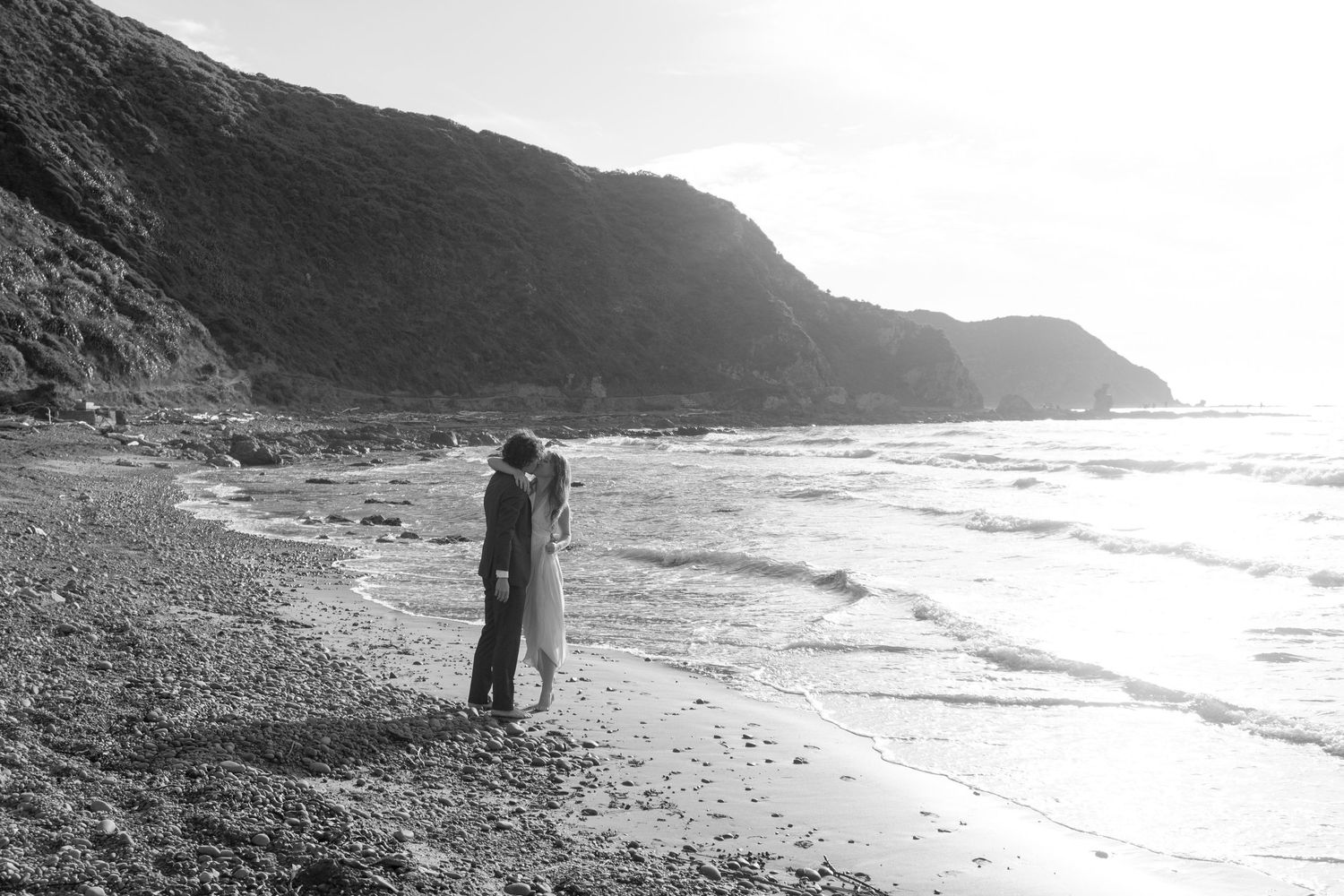 Black and white photo of a couple embracing on a rocky beach with dramatic cliffs and ocean waves in the background.