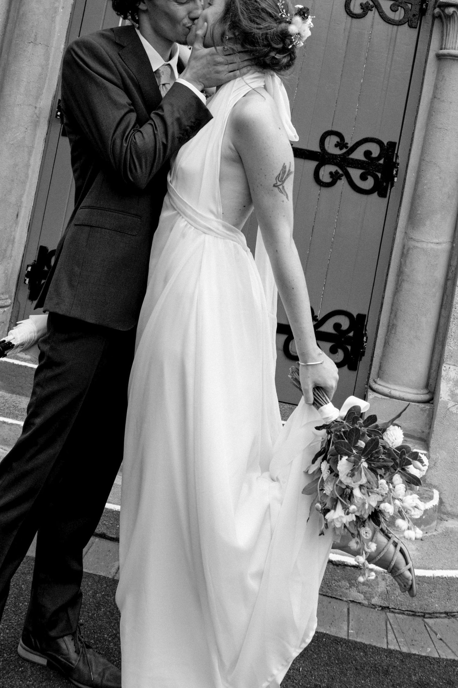 Black and white photo of a bride and groom sharing a kiss outside a church, bride holding a bouquet.