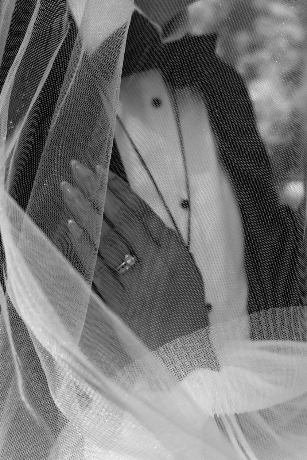 Black and white wedding photo of bride's hand with engagement ring resting on groom's chest, framed by veil.