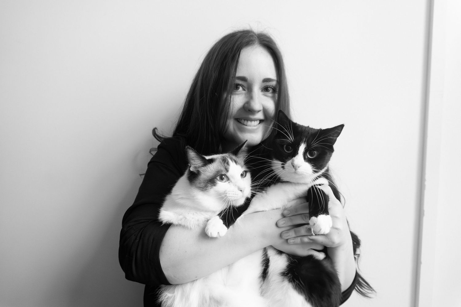 Black and white photo of smiling woman holding two cats, a tuxedo and a ragdoll, against a light wall.