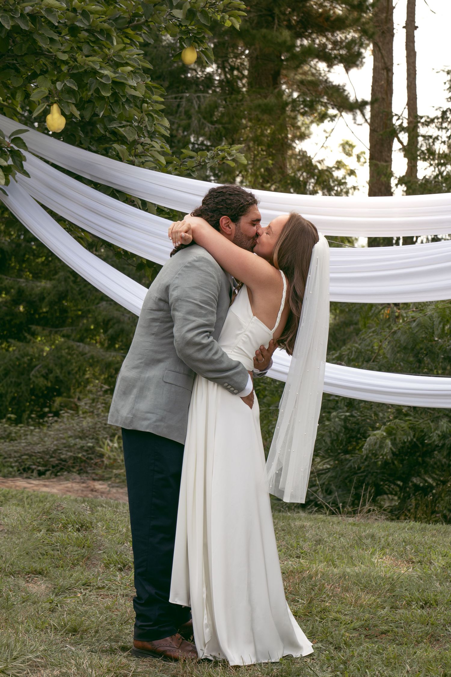 Bride and groom share first kiss at outdoor wedding ceremony with white draped fabric backdrop.