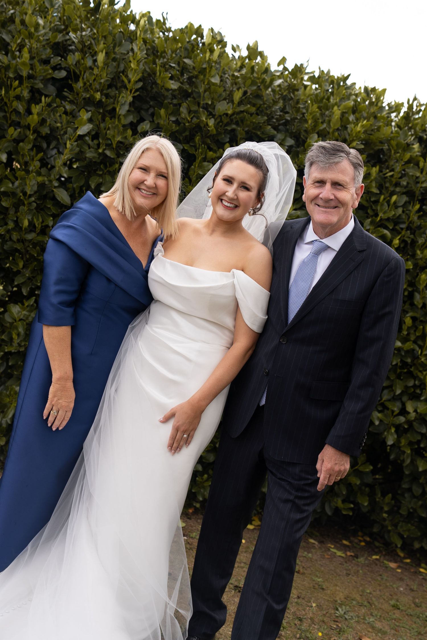 Bride in white off-shoulder gown with veil poses with parents in formal attire before green hedge backdrop.