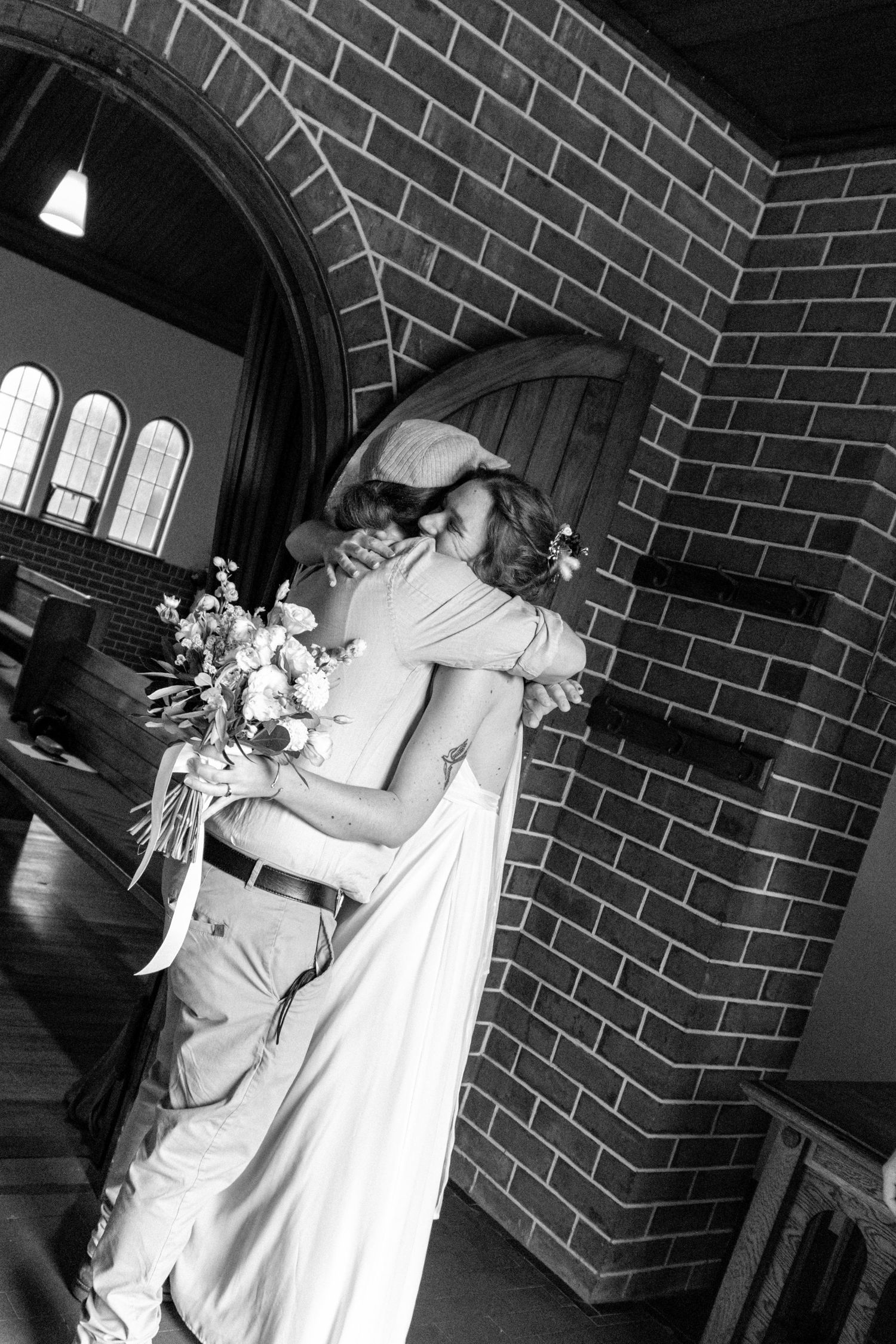Black and white photo of a bride and groom embracing in a brick church, bride holding a floral bouquet.