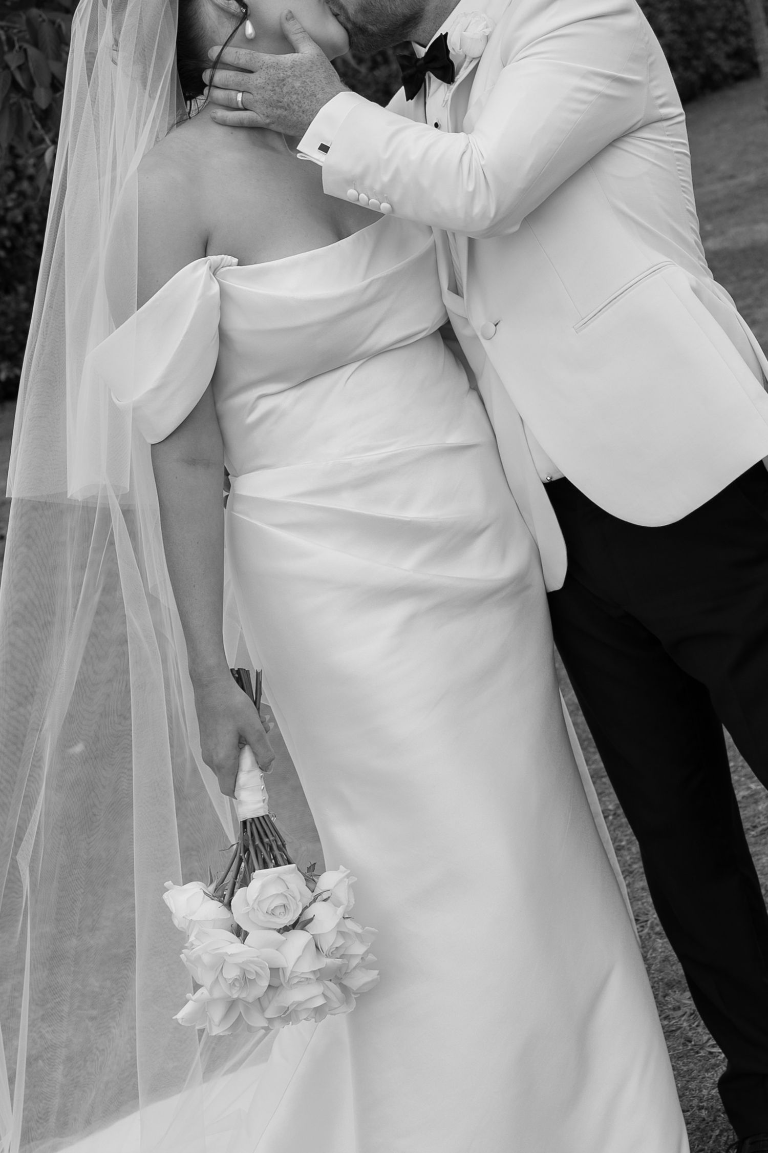 Black and white photo of bride in strapless gown holding rose bouquet, groom in white tuxedo embracing her.
