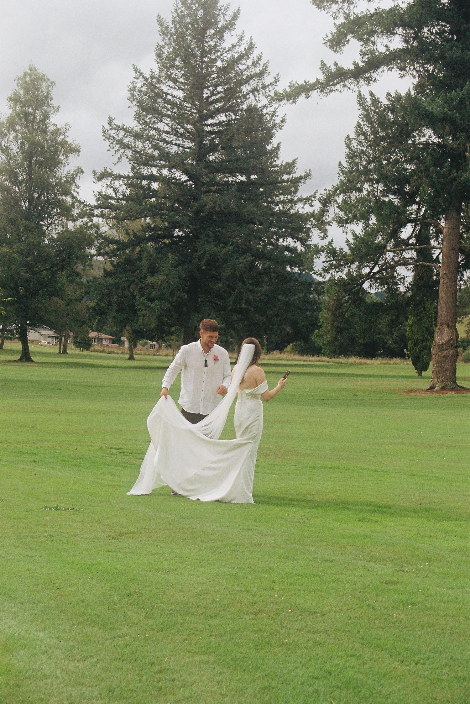 Bride and groom walking on lush green lawn, bride's white gown flowing in wind, tall evergreen trees in background.