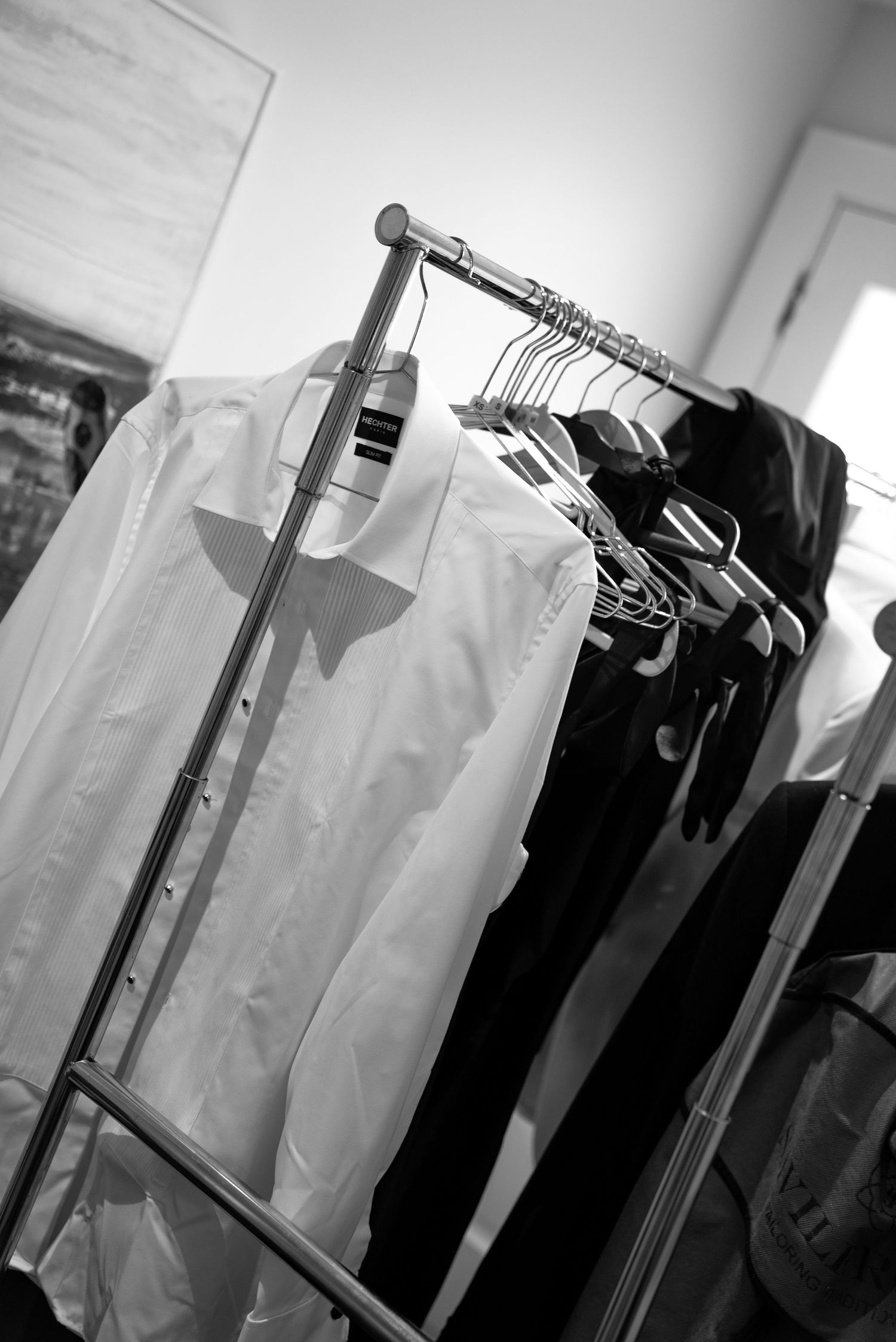 Black and white photo of dress shirts and dark clothing hanging neatly on a metal clothing rack indoors.