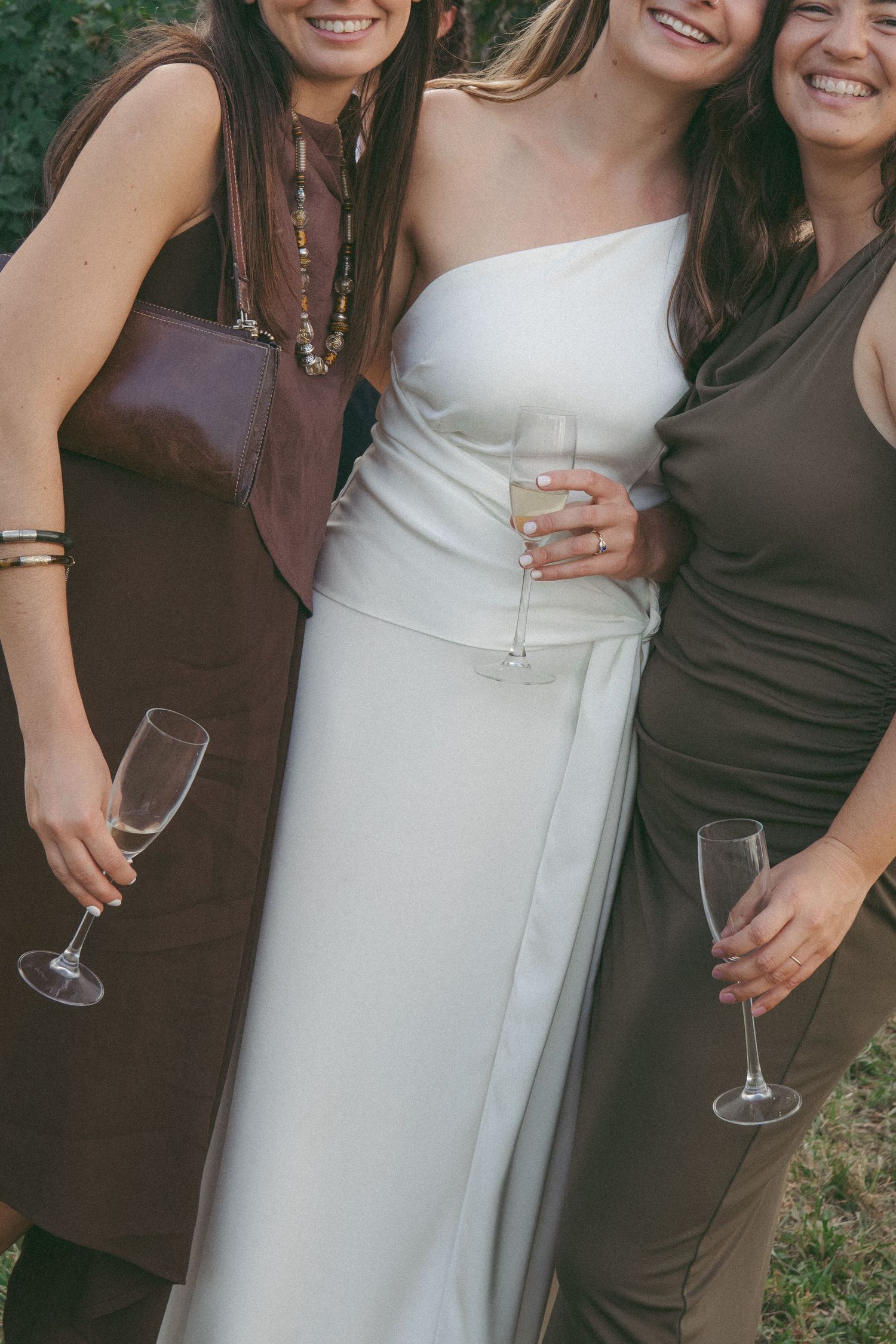 Three women in brown and white dresses smiling and holding champagne flutes at a wedding celebration outdoors.